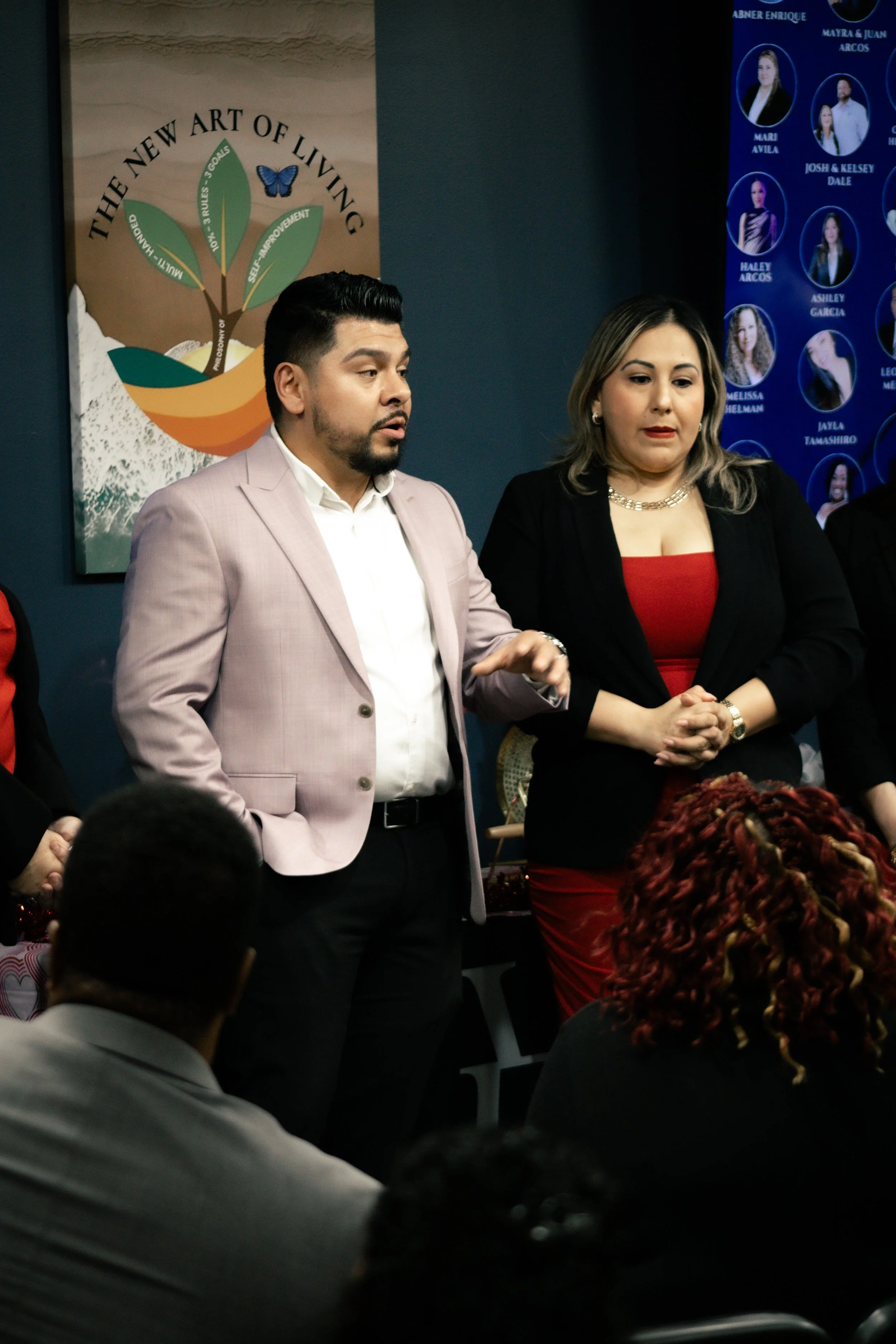 A man in a light pink blazer and a woman in a red dress and black blazer standing in front of an audience at a conference or meeting. Behind them are posters and a blue wall.