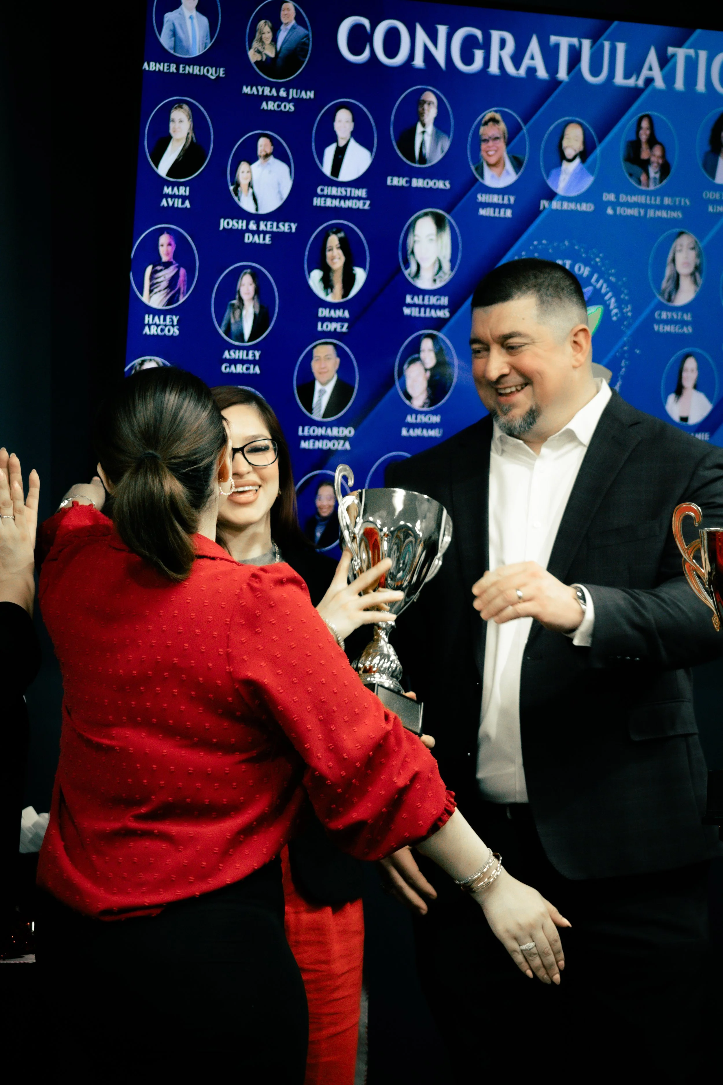 A woman in a red blouse receiving a trophy from a man in a black suit at a celebration event. A blue background with headshots and names of various people is visible behind them.