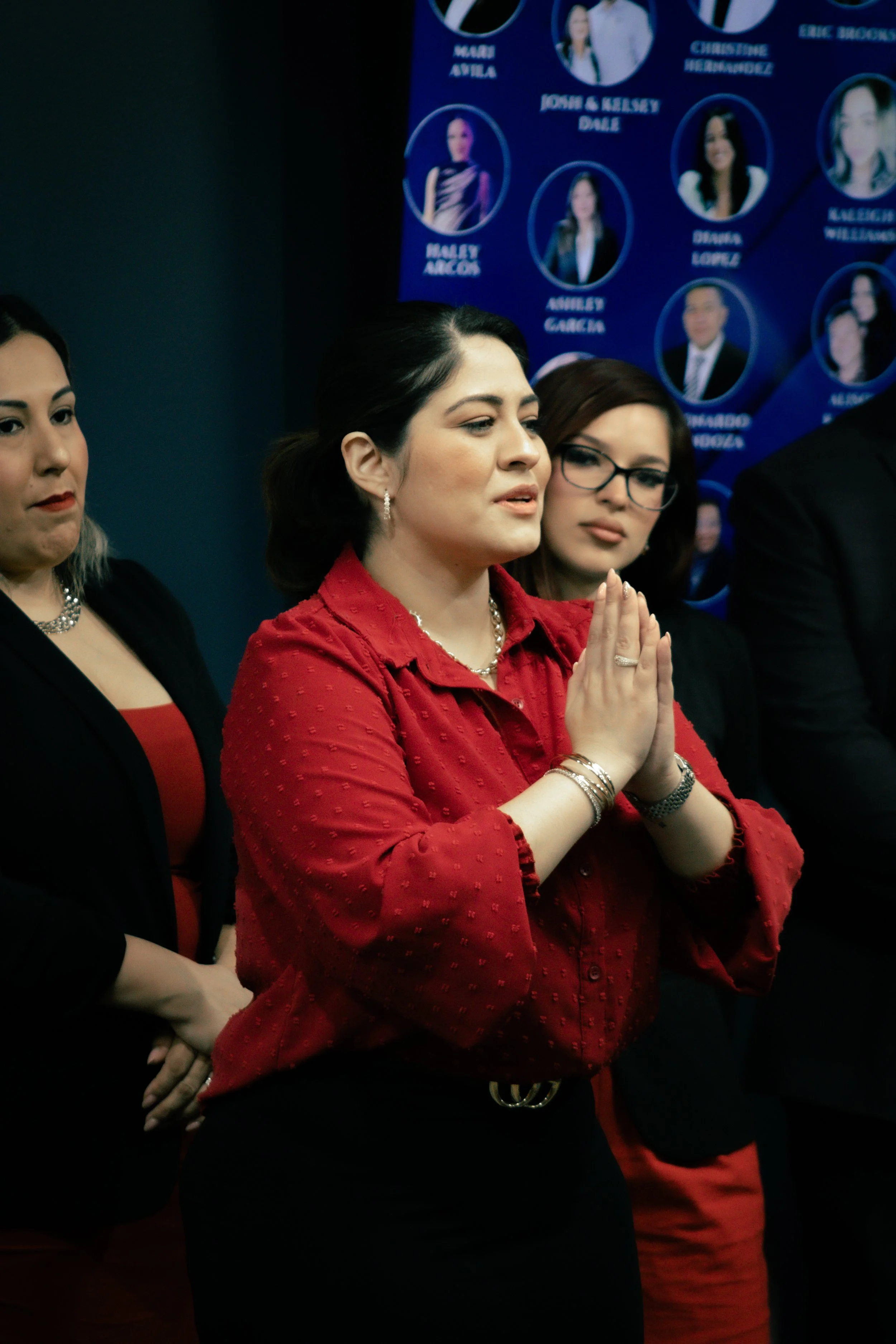 A woman in a red blouse standing with her hands pressed together in front of her chest during a formal event, with other women and a blue background with photos of people in circles.