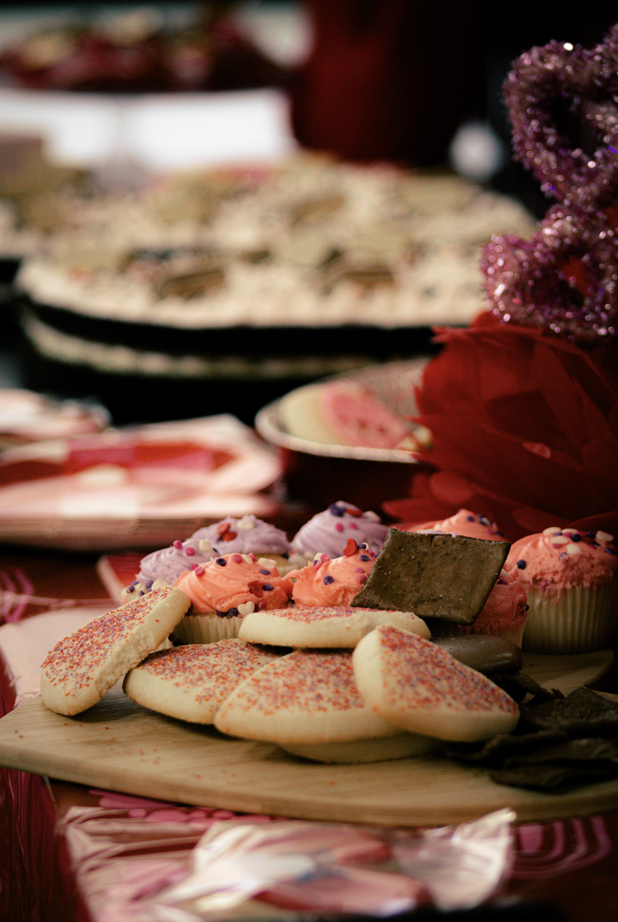 Assorted Valentine's Day cookies and cupcakes on a wooden platter, with pink and purple decorations in the background.