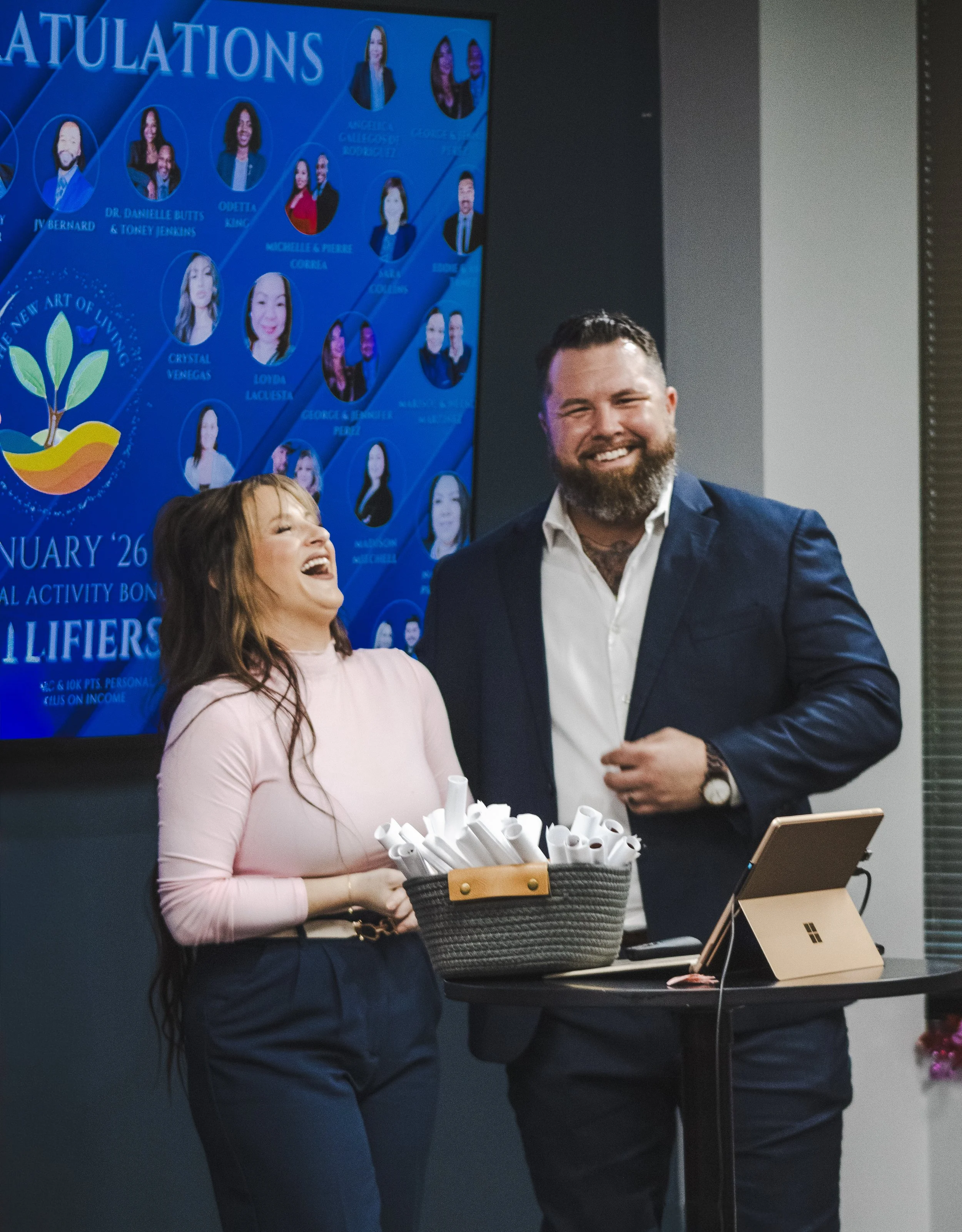 Two people, a woman and a man, are smiling and laughing at an event. The woman has long brown hair and is wearing a light pink top and dark pants. The man has a beard, short hair, and is in a dark suit with an open-collared white shirt. They are stan