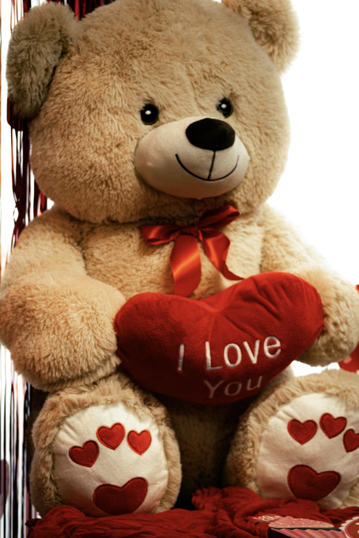 A large, plush teddy bear holding a red heart pillow with the words 'I Love You' embroidered on it. The bear has beige fur, a smiling face, and a red satin ribbon around its neck. Its paws are decorated with red hearts.
