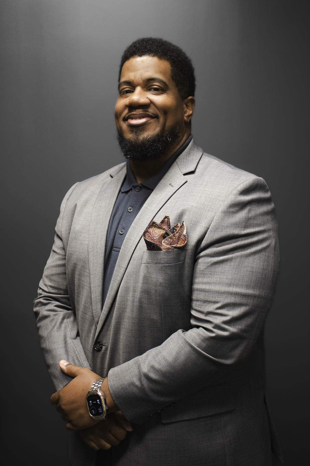 A professional man in a gray suit and dark shirt standing against a dark background, smiling and looking at the camera, with a watch on his wrist and a pocket square in his suit pocket.