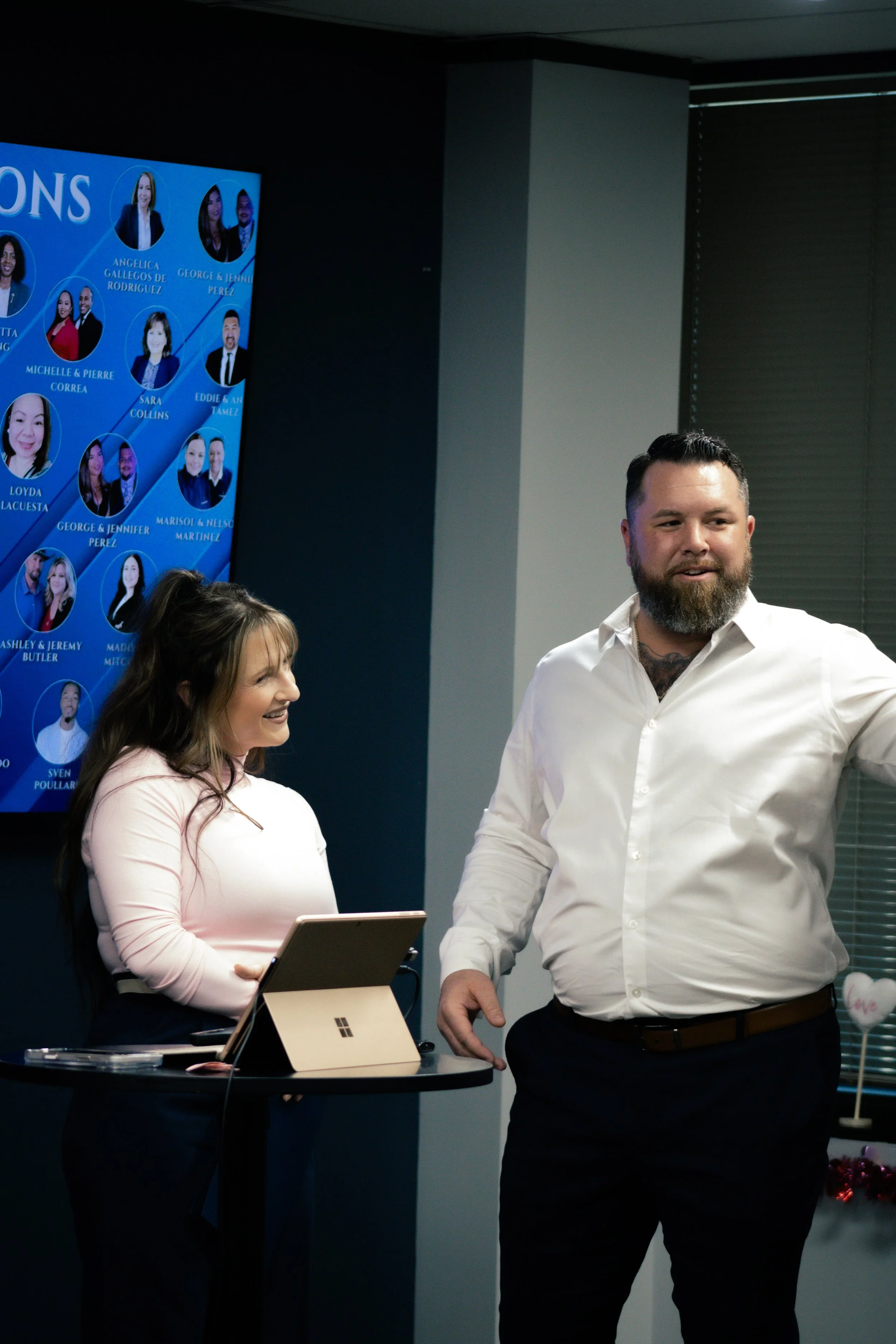 Two people standing in front of a presentation screen with a virtual campaign plan, including headshots and names of team members, in a modern office setting.