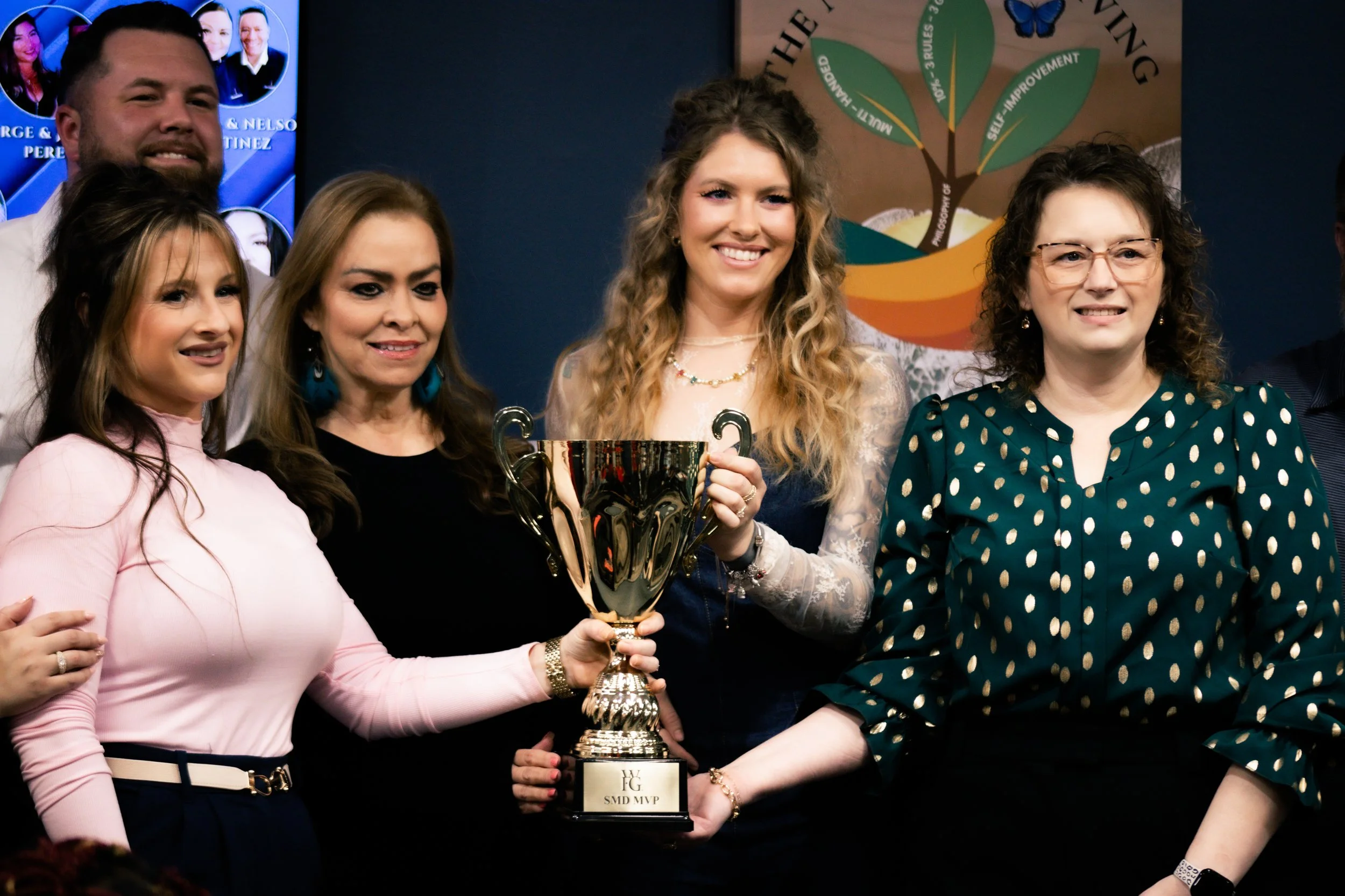 Group of six women and one man at an award celebration. One woman is holding a large gold trophy labeled 'SMD MVP' and is smiling for the photo. They are standing in front of a colorful poster.