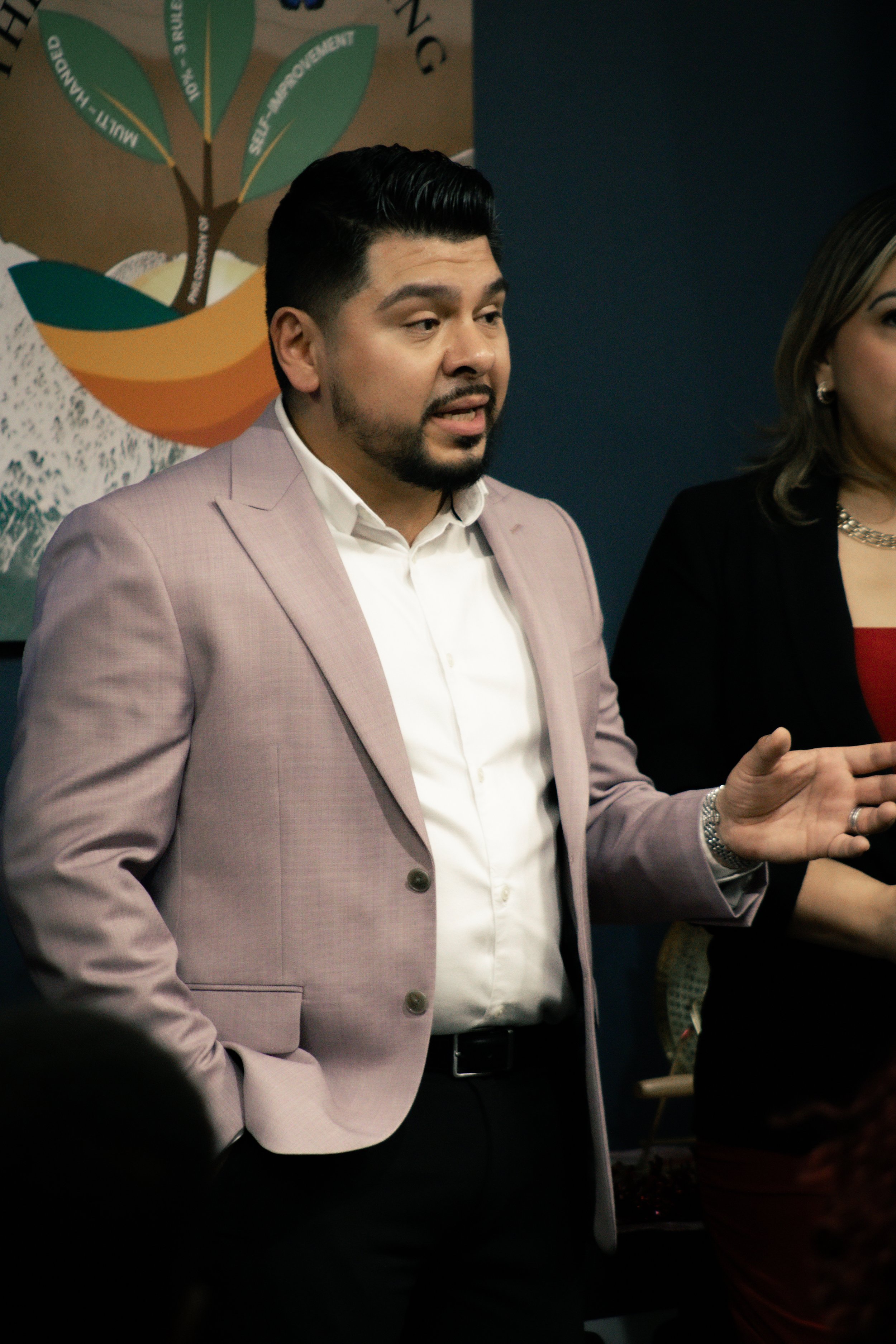 A man in a light pink suit and white shirt speaking, with a woman standing beside him, in a professional setting.