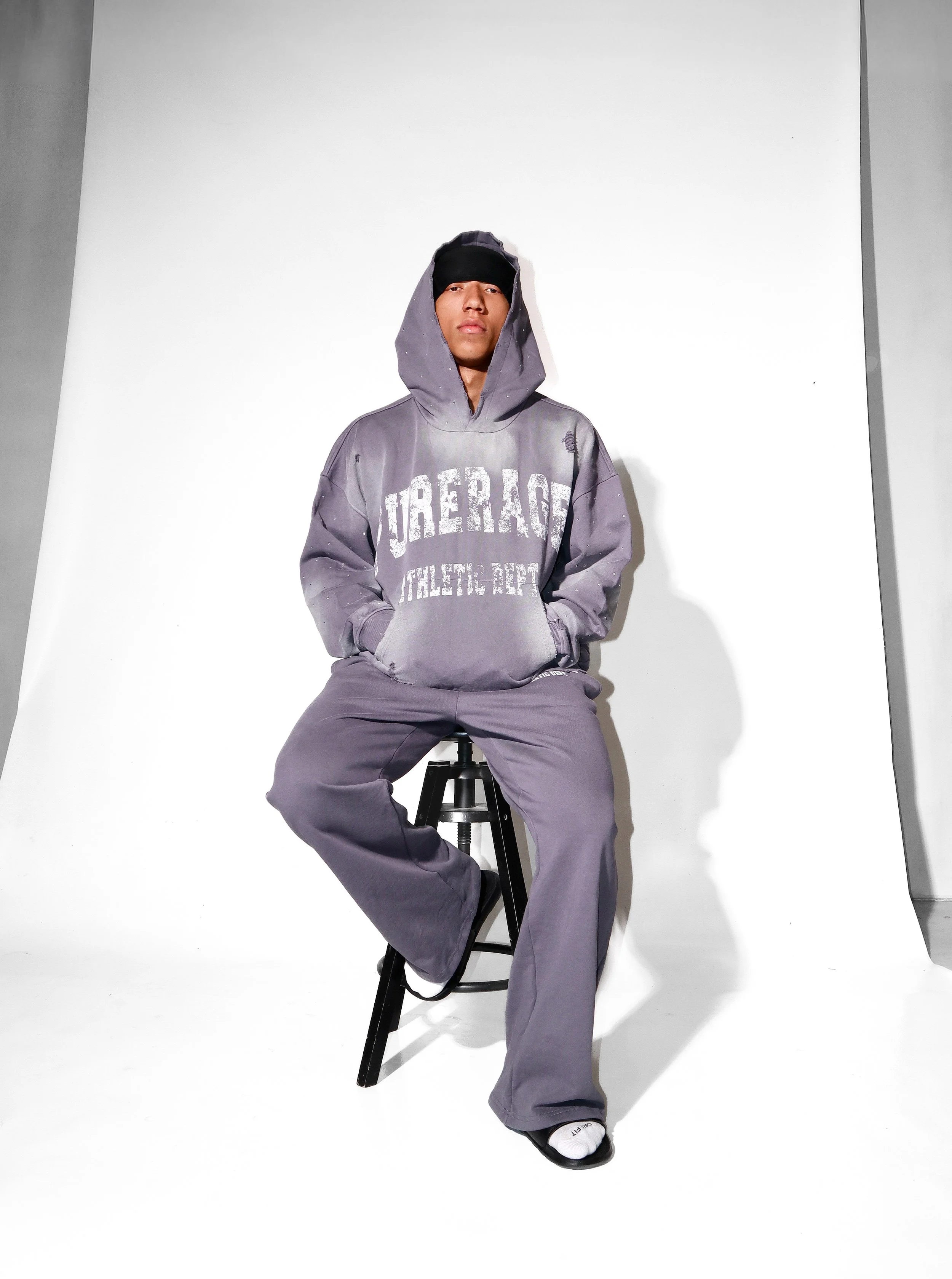 A young man in a gray hoodie and sweatpants sitting on a black stool in a photography studio with white walls, wearing a black headband and white socks.