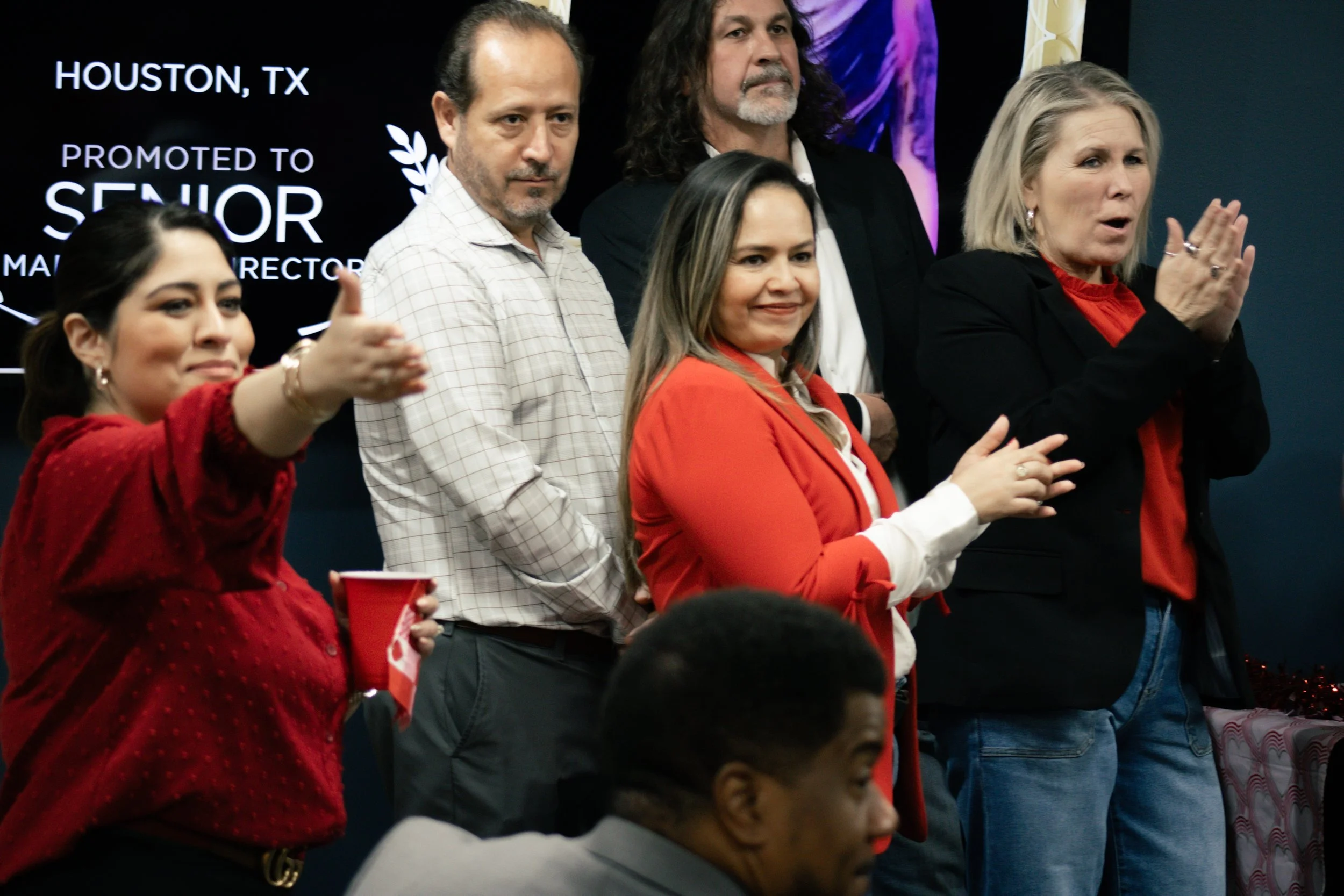 Group of people standing on stage in front of a black screen with white text, celebrating in Houston, TX. One woman is giving a thumbs up and holding a red cup, others are smiling and clapping.