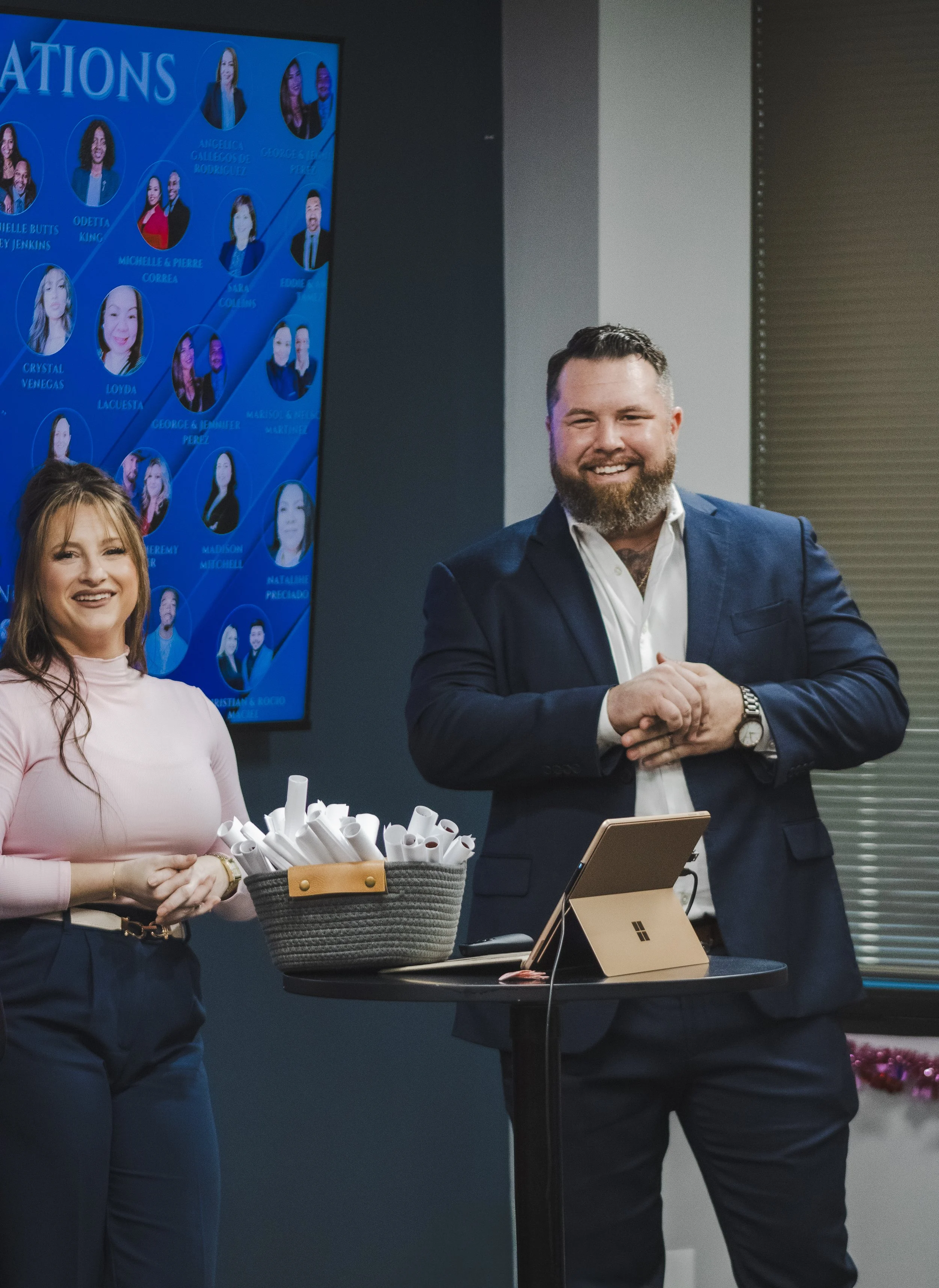 Two smiling professionals, a woman in a pink turtleneck and dark pants and a man in a navy blazer and white shirt, standing in a modern office with a screen displaying corporate headshots and names in the background. A table holds a basket of rolled 
