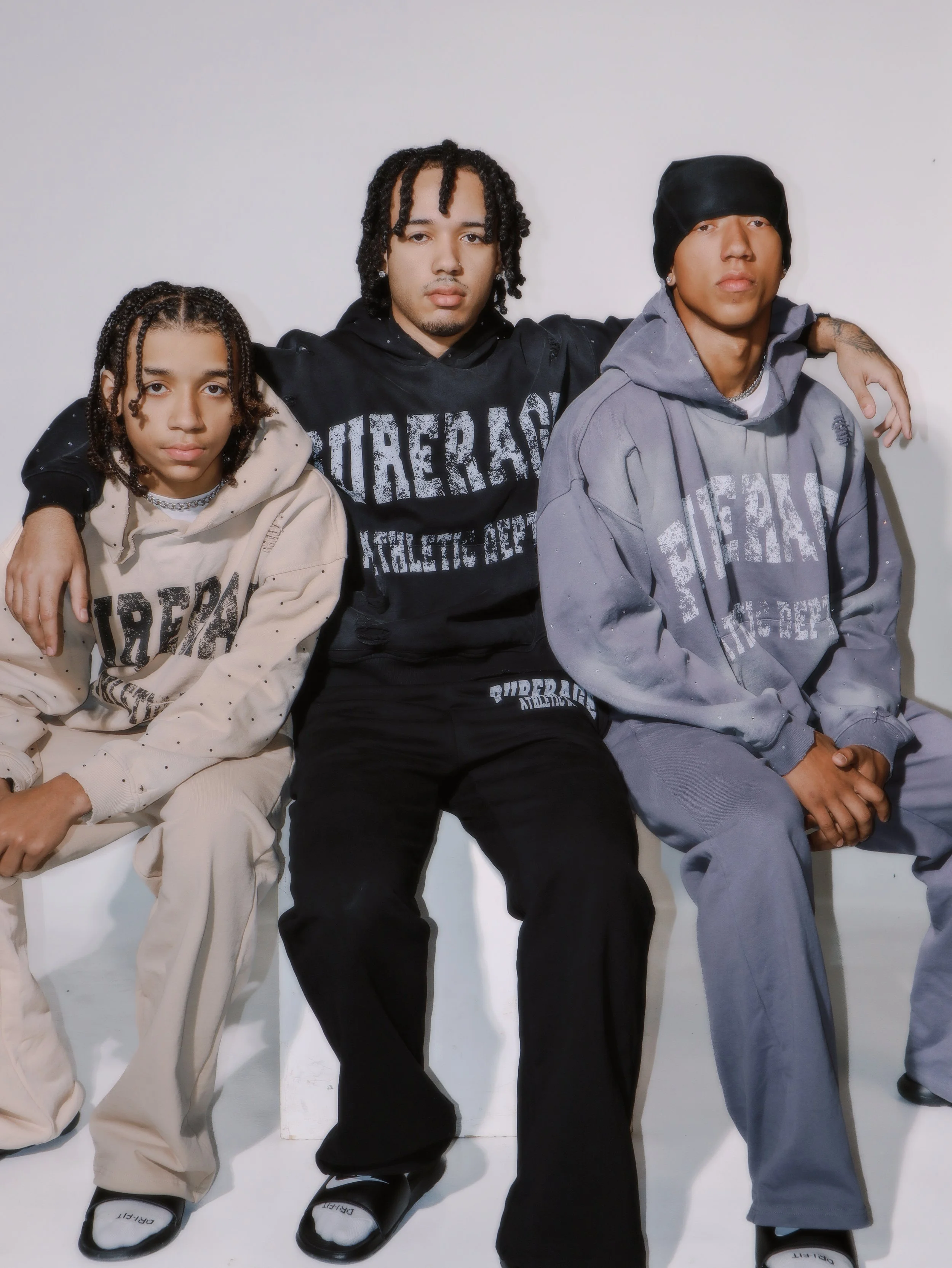 Three young men sitting together against a white background, wearing hoodies with the word 'pureage' on them in different colors.