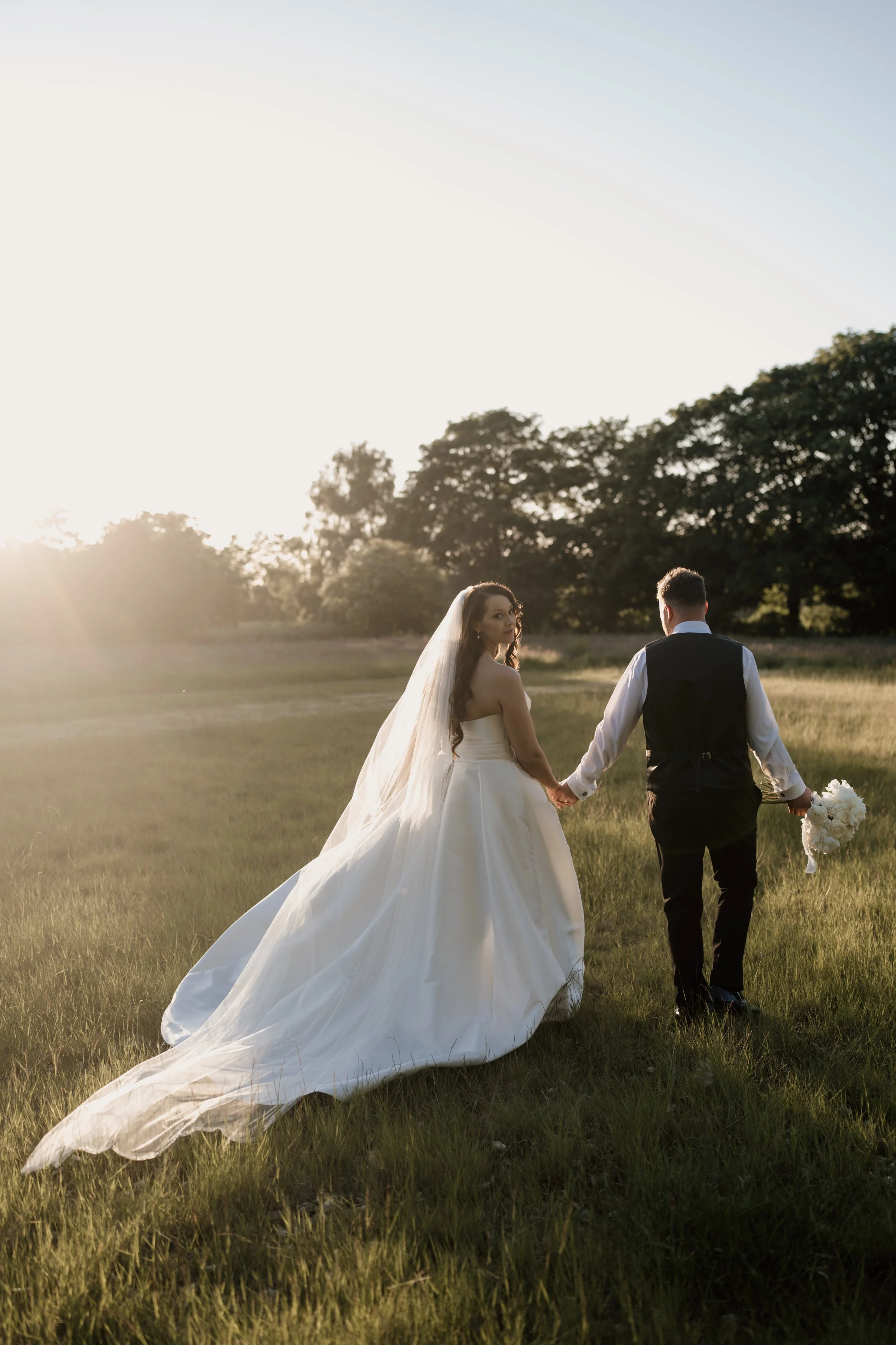Bridal couple walking hand in hand in a grassy field during sunset, with the bride in a white wedding gown and veil, and the groom in a dark vest and white shirt, holding a bouquet.