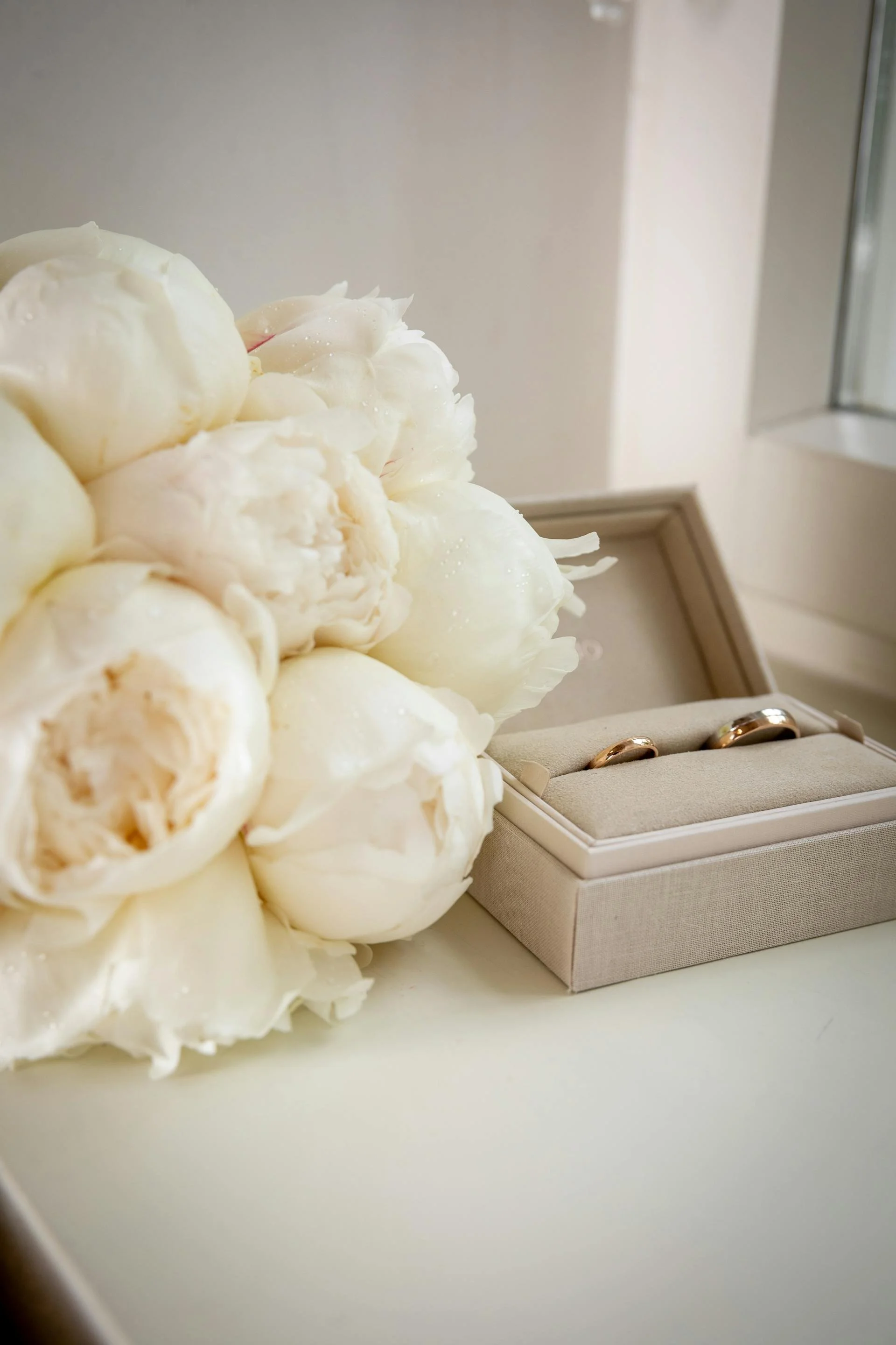 Close-up of white flowers and a jewelry box with two wedding rings on a white surface.