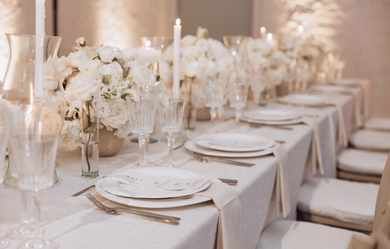 Elegant wedding or event table setup with white floral arrangements, candles, and glassware on a long white table with beige chairs.