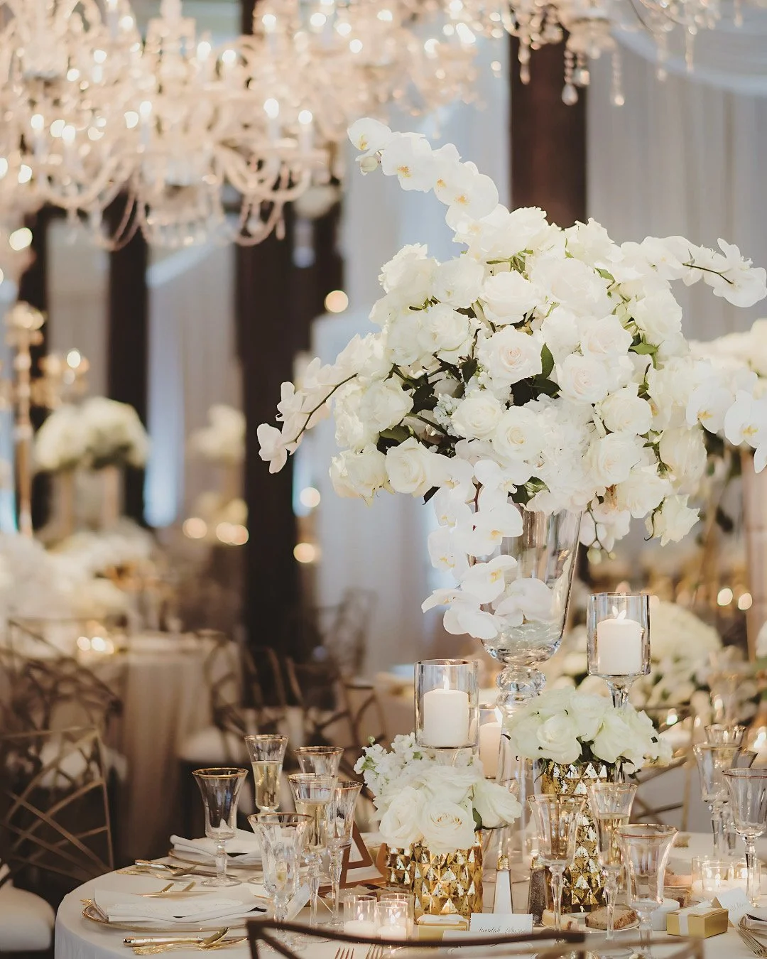 Elegant wedding reception table with large floral arrangements of white roses and orchids, surrounded by glass candle holders and chandeliers in the background.