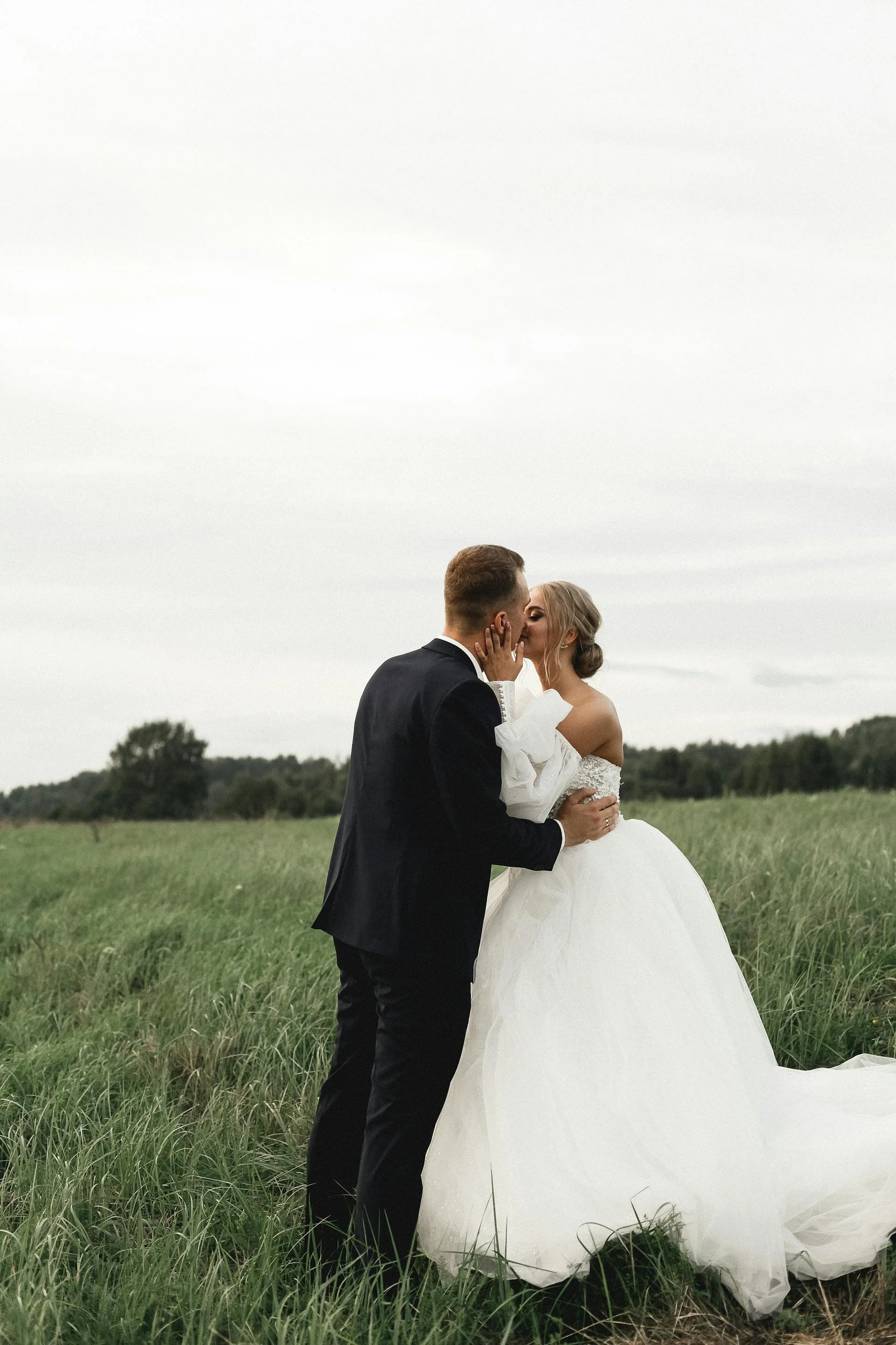 Bride and groom sharing a kiss in a grassy field, bride in a white wedding gown and groom in a black tuxedo, overcast sky.