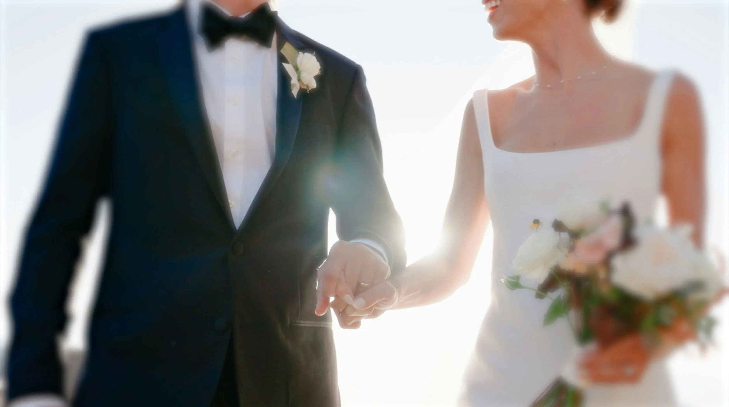 A bride and groom holding hands during a wedding ceremony, with the bride in a white dress and holding a bouquet, and the groom in a black suit with a bow tie, against a bright background.
