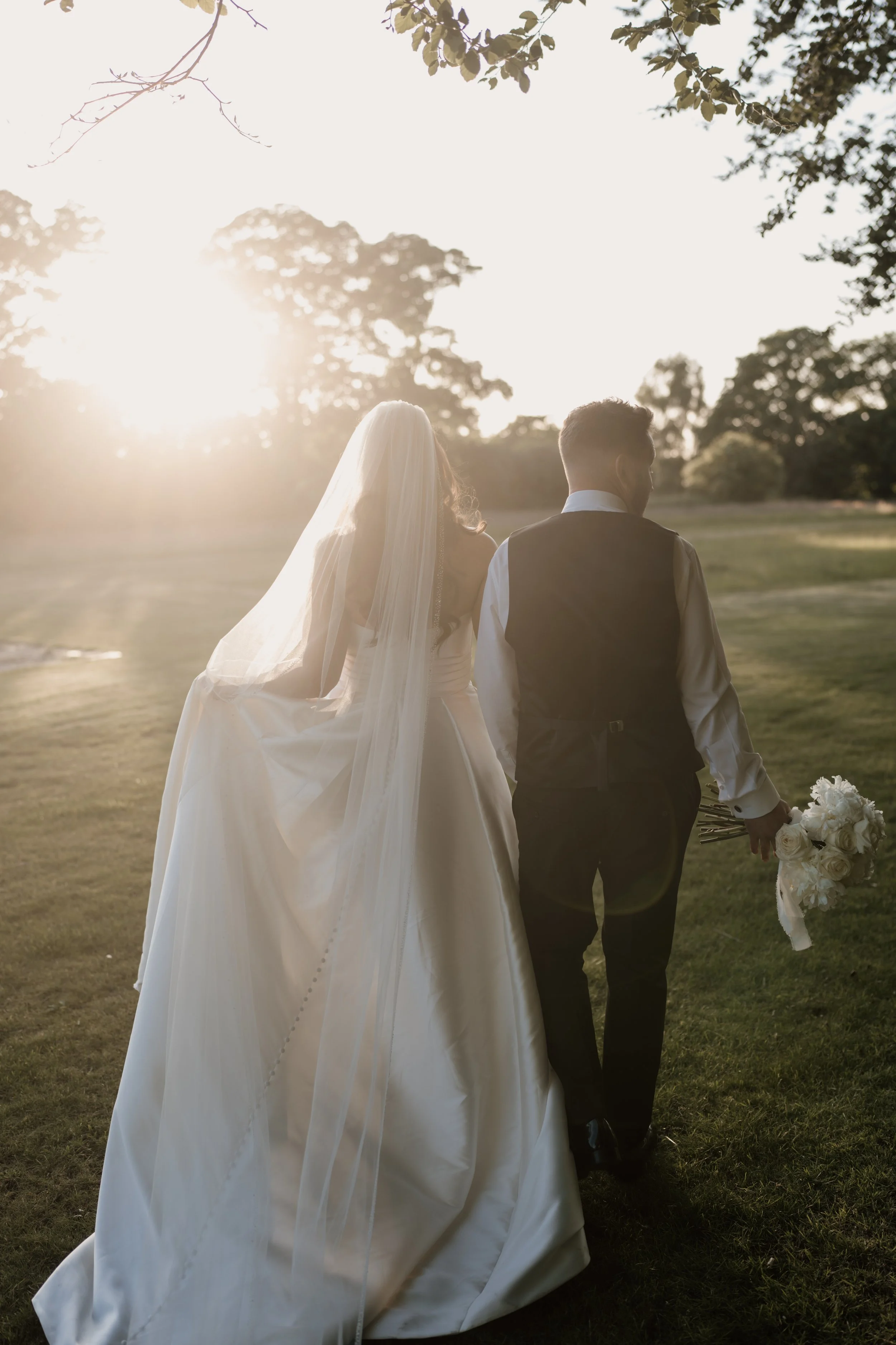 Bride and groom walking outdoors at sunset, the bride in a white gown with veil holding a bouquet, the groom in dark suit with a white shirt.