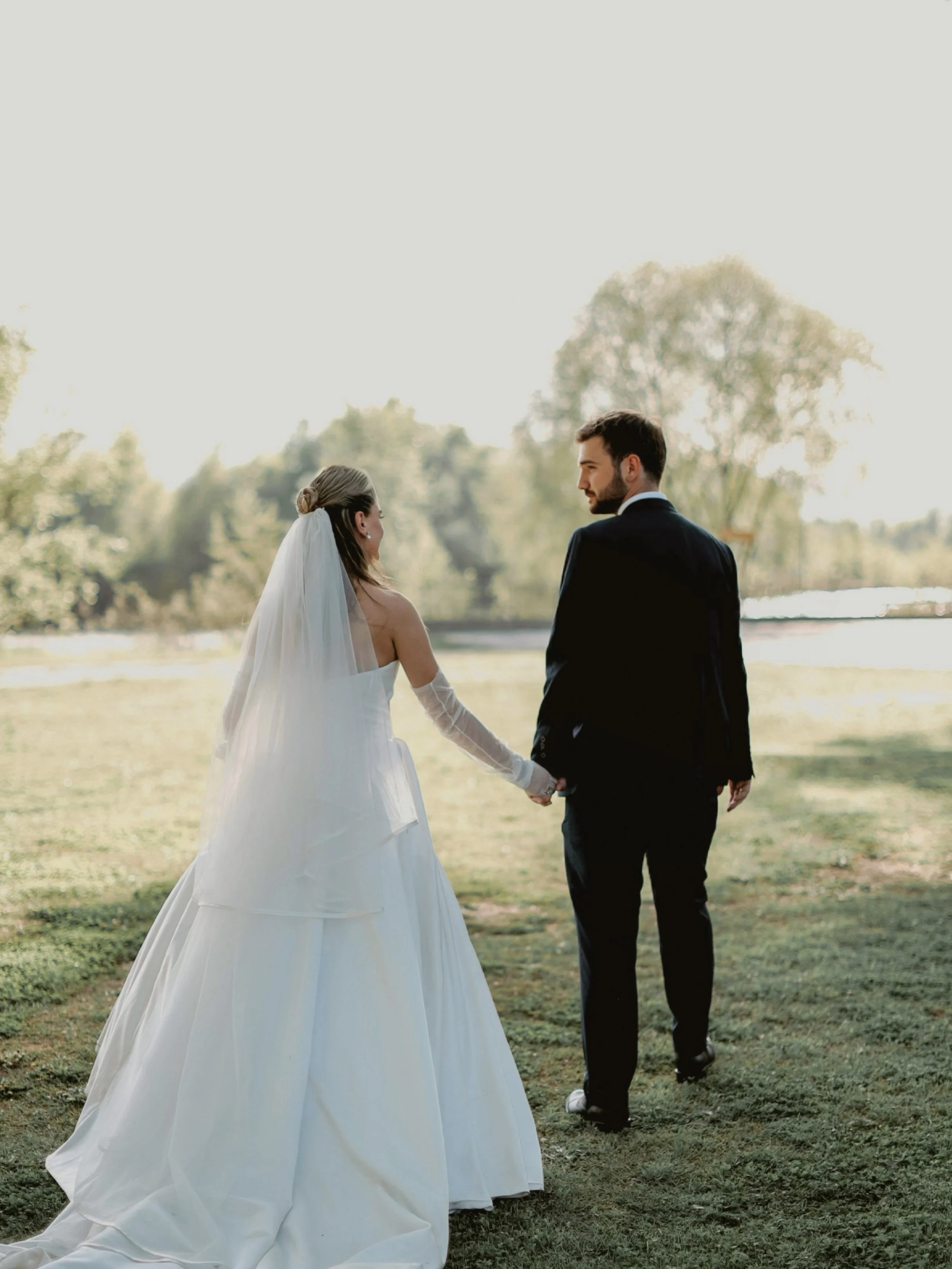 A bride and groom holding hands while walking outdoors in a grassy area with trees in the background, during a wedding ceremony.