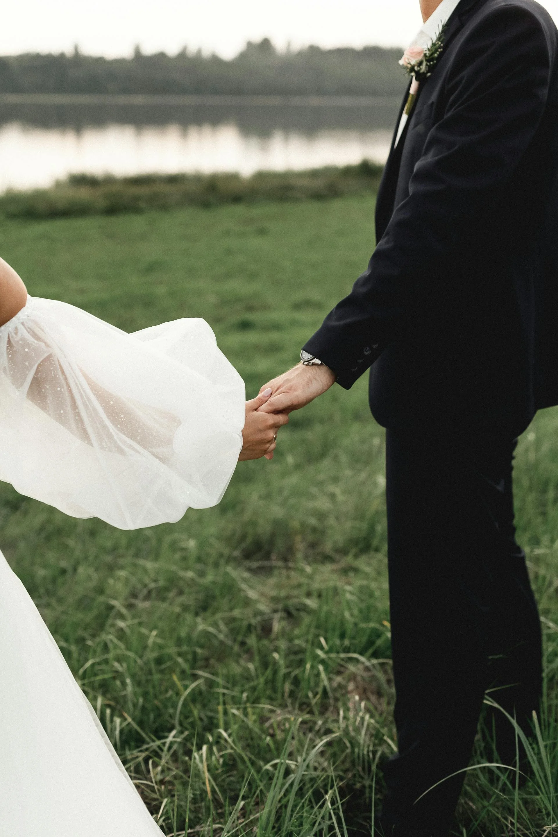 A couple holding hands outdoors near a body of water on their wedding day. The bride is in a white dress with sheer puffed sleeves, and the groom is in a dark suit with a boutonniere. The background shows a grassy area and a lake with trees in the distance.