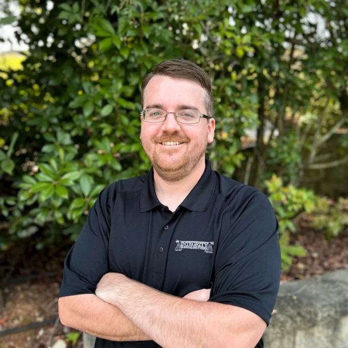 a smiling man with folded arms and glasses and scruff in front of a green tree