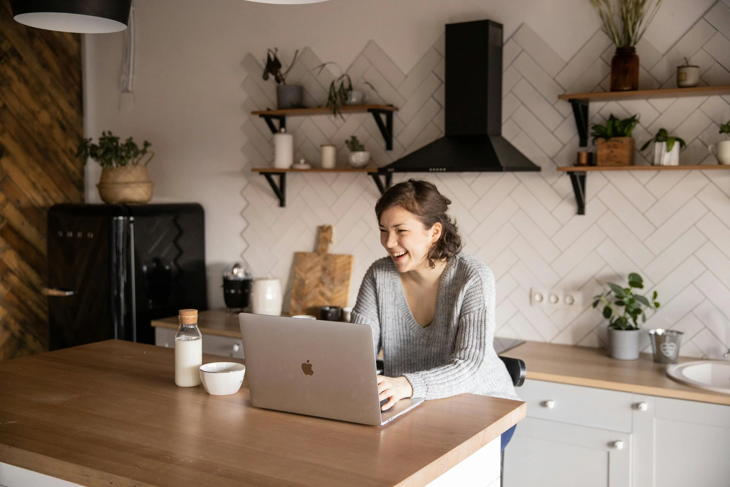 A young woman with short dark hair, wearing a grey sweater, sitting at a wooden kitchen counter, smiling and looking at her open MacBook.