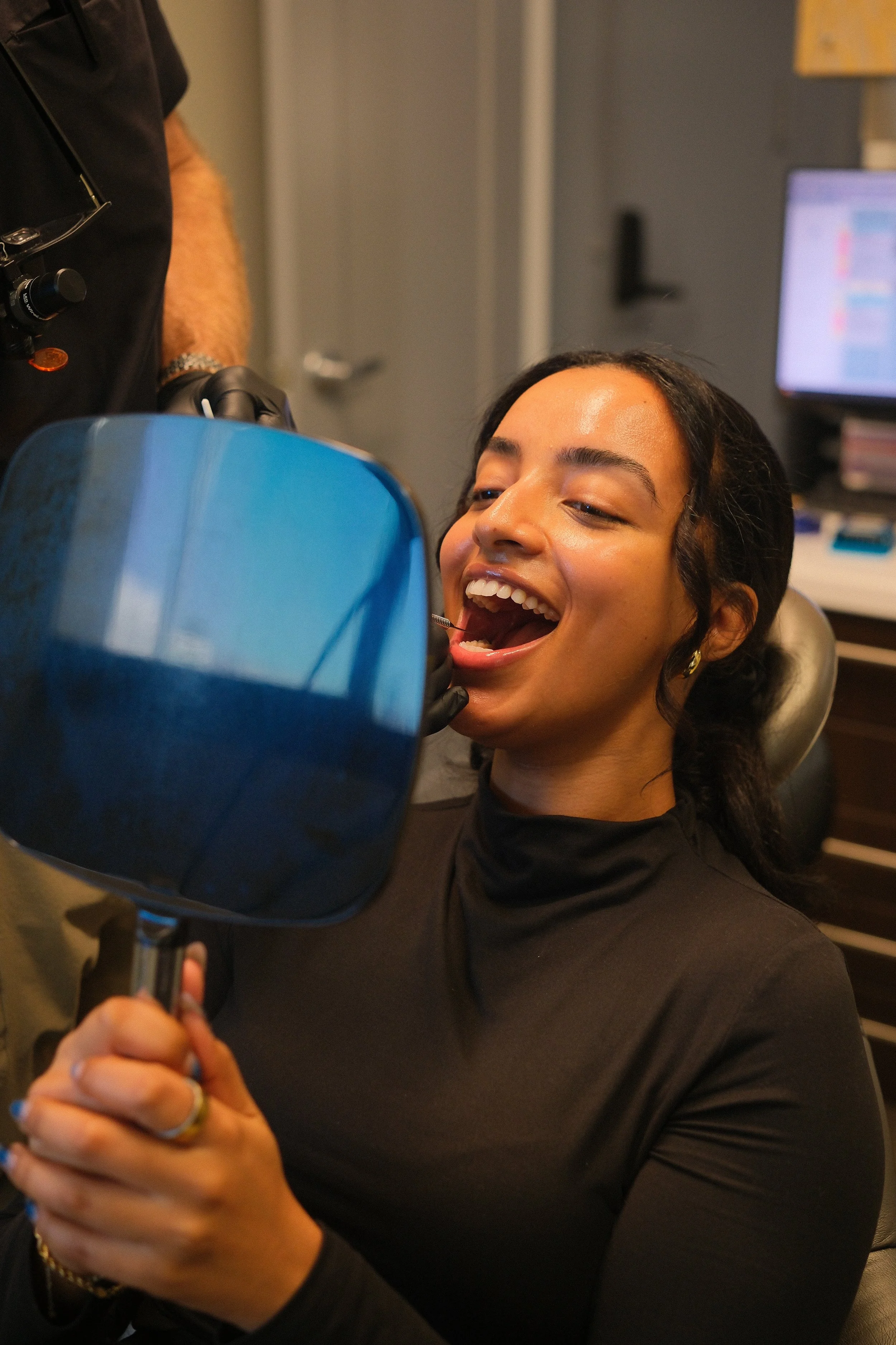 A smiling woman in a dental chair holding a red mirror, with dental bibs around her neck, in a dental office setting.