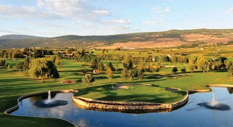 A scenic view of Okanagan Golf Club in Kelowna featuring a beautifully manicured island style green surrounded by water, lush fairways and rolling Okanagan hills in the background.