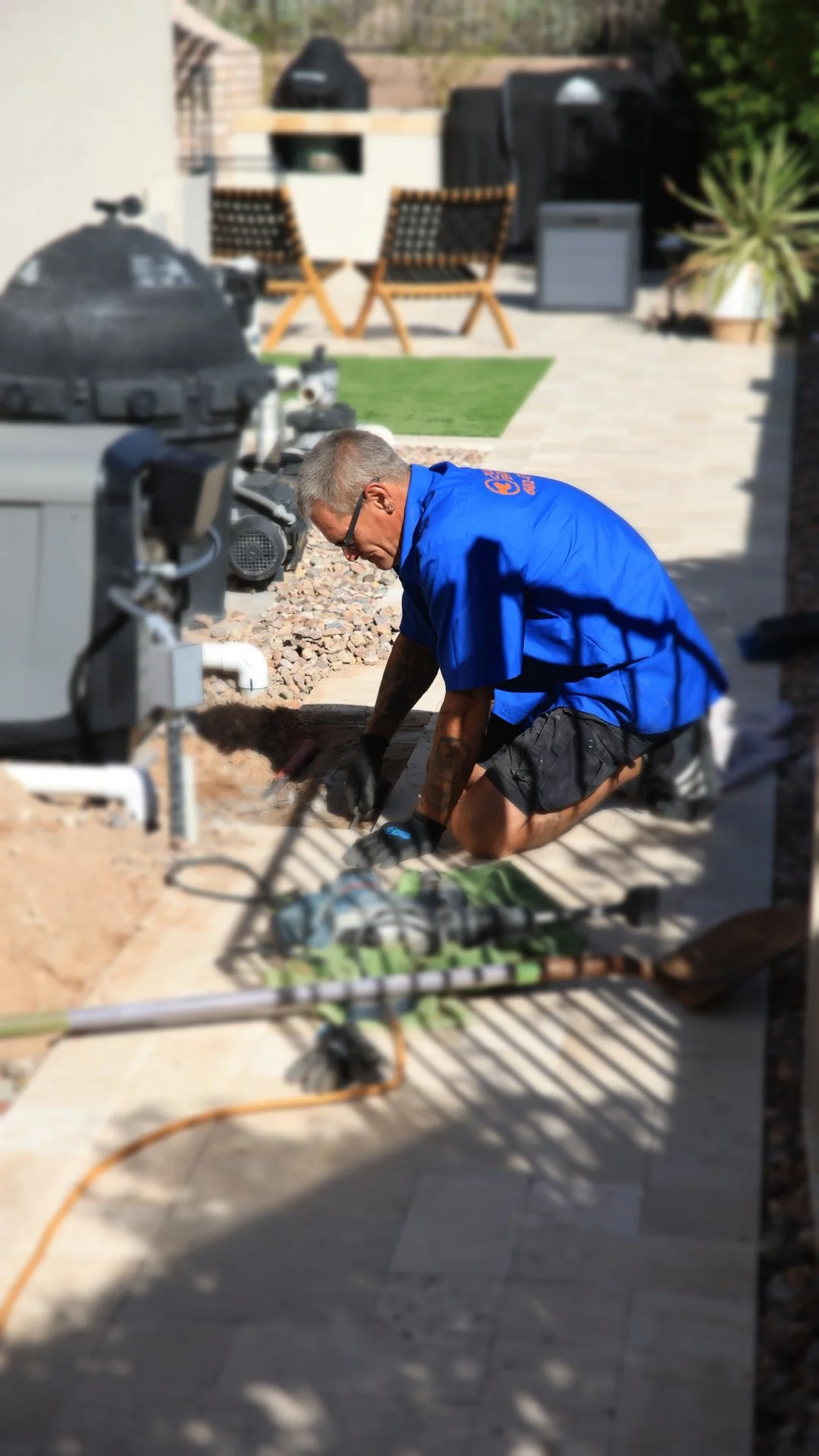 A plumber kneeling on a backyard patio working on underground  pipe system, surrounded by construction tools and equipment.