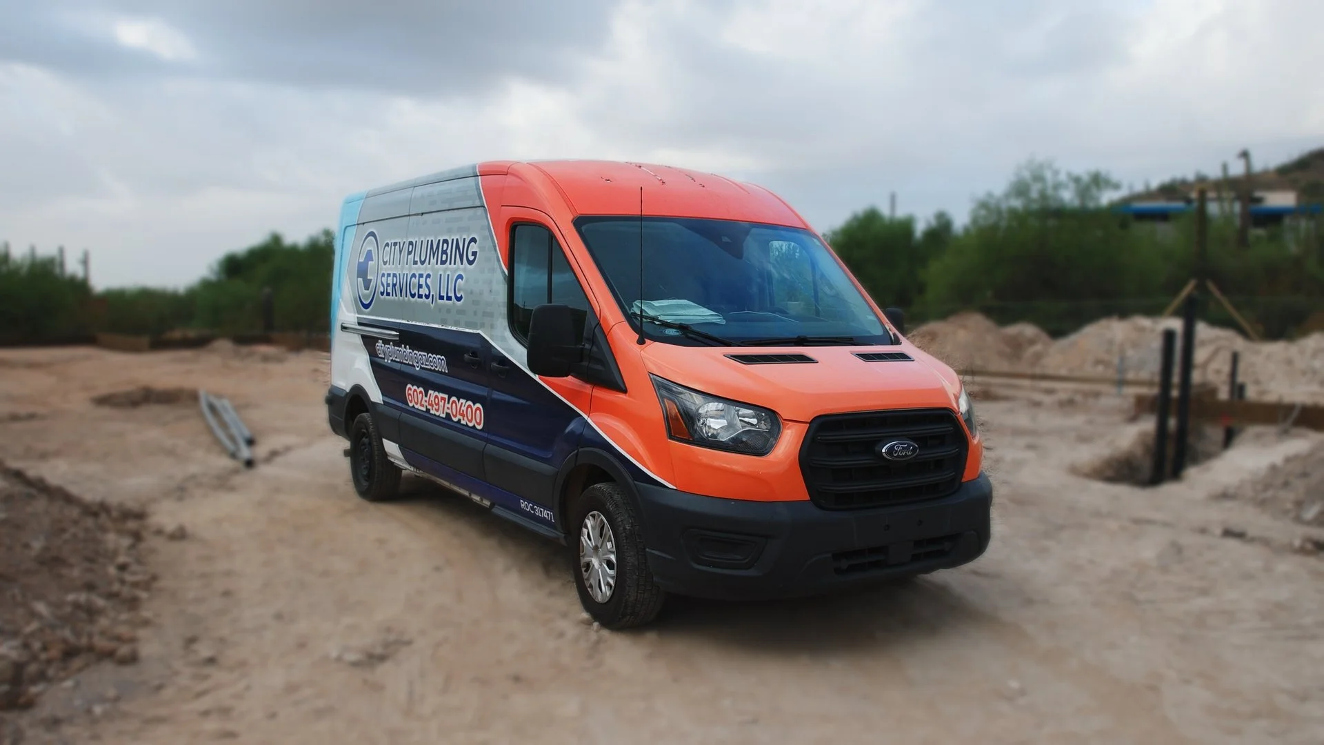 A orange and white commercial van with the logo and name of City Plumbing Services, LLC, parked on a construction site with dirt and some greenery in the background.