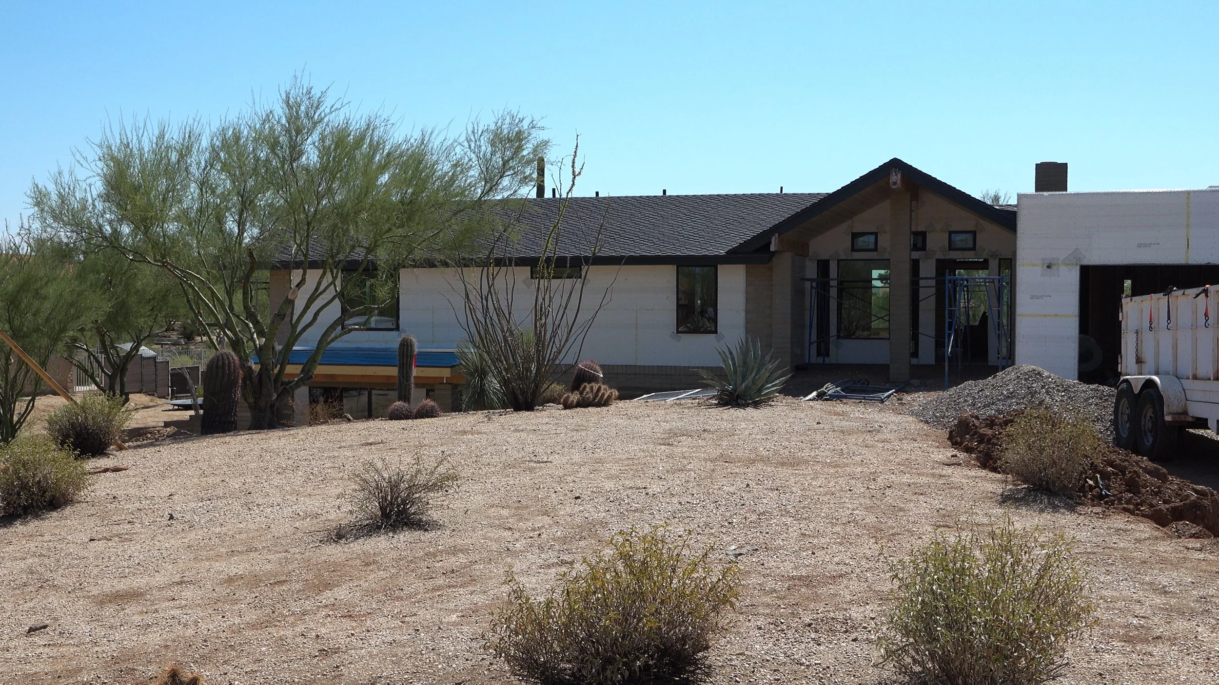 A house under construction in a desert landscape with sparse bushes, cacti, and a large tree, a trailer on the right, and a clear blue sky.