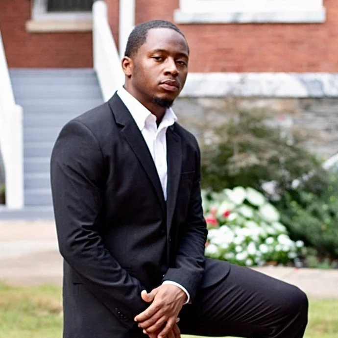 A young man in a black suit and white shirt posing outdoors in front of a brick house with flowers and greenery.