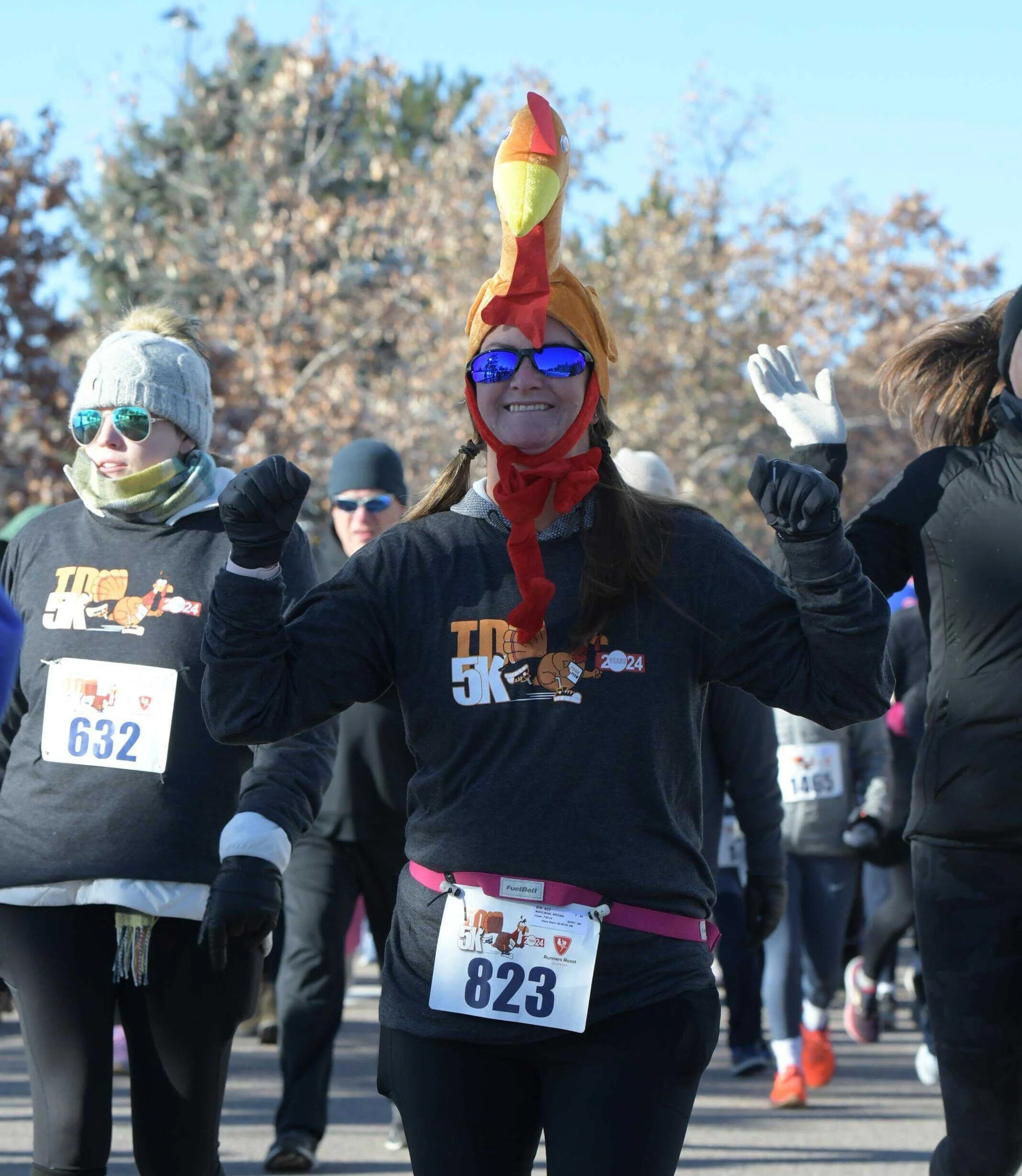 A woman with a turkey hat and sunglasses celebrating at the Turkey Day 5K - Highlands Ranch, surrounded by other runners in warm clothing.