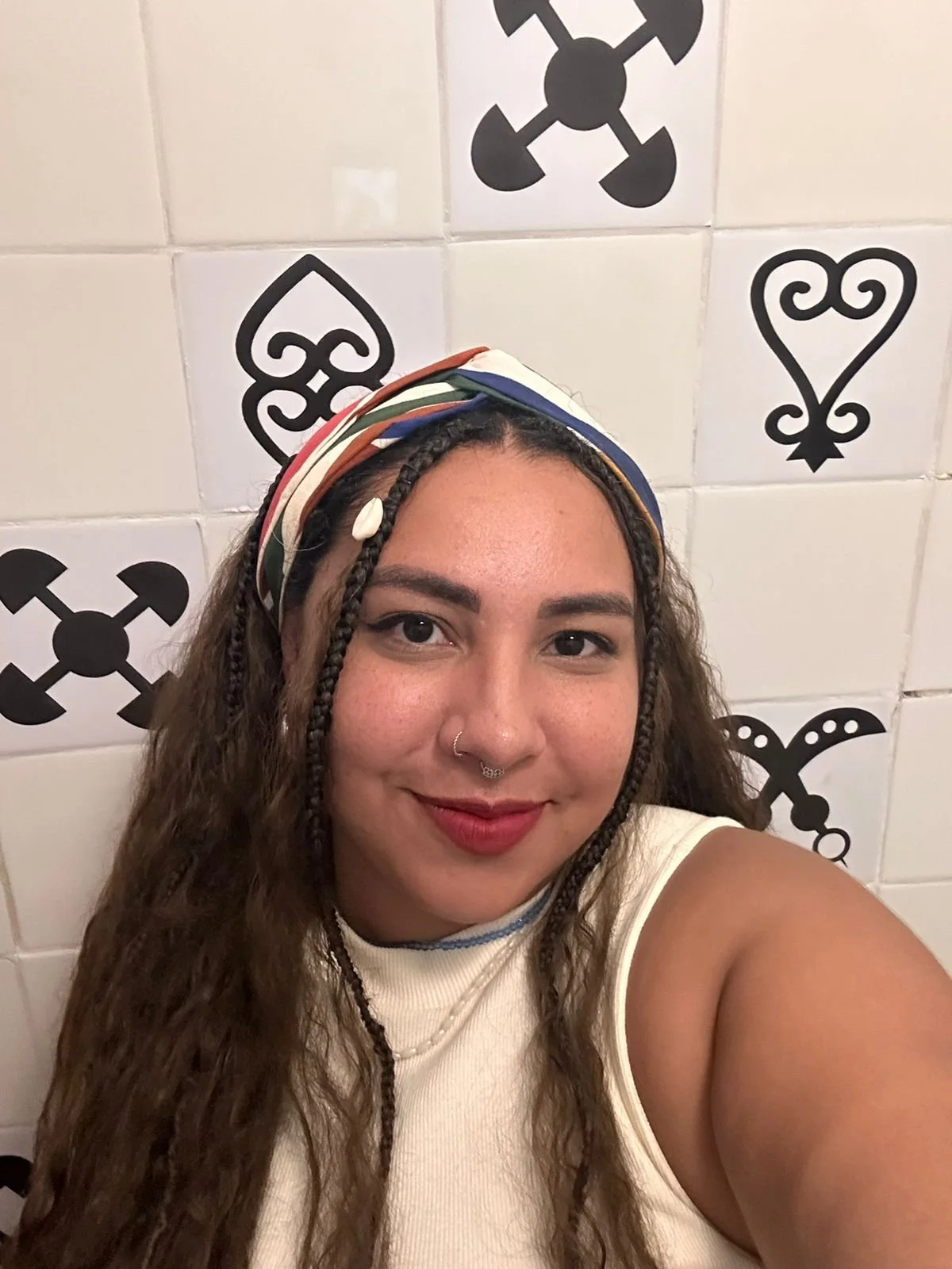 Mulher sorrindo para a câmera, usando lenço colorido na cabeça, com cabelo cacheado, ao fundo parede com azulejos brancos decorados com símbolos pretos.