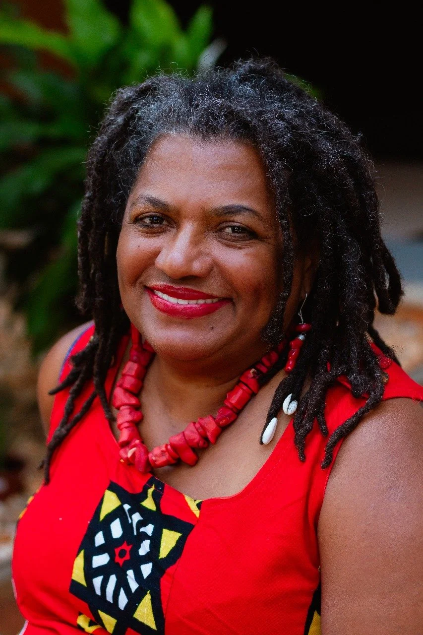 Mulher negra com dreadlocks, vestindo roupa vermelha com padrão africano, usando colar de contas vermelhas e brincos brancos, sorrindo em ambiente ao ar livre com vegetação ao fundo.