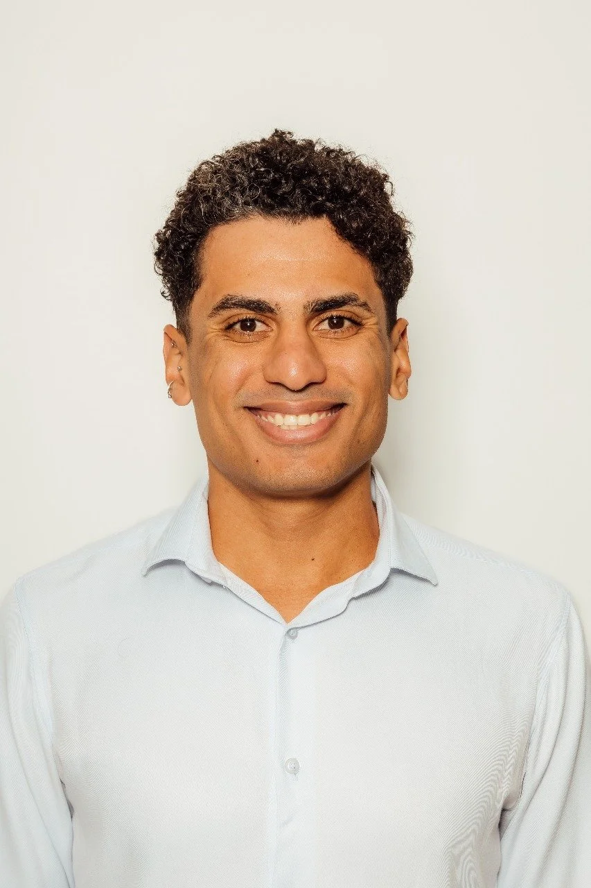 Homem sorridente com cabelo cacheado, usando camisa social branca, contra fundo branco.
