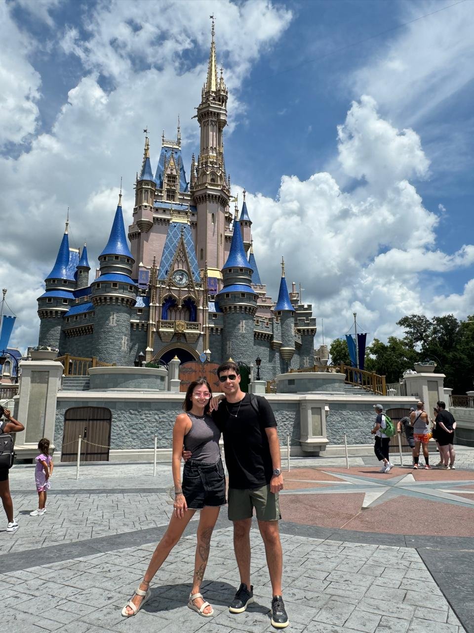 Casal em frente ao castelo da Disney, com nuvens no céu e outras pessoas ao redor.