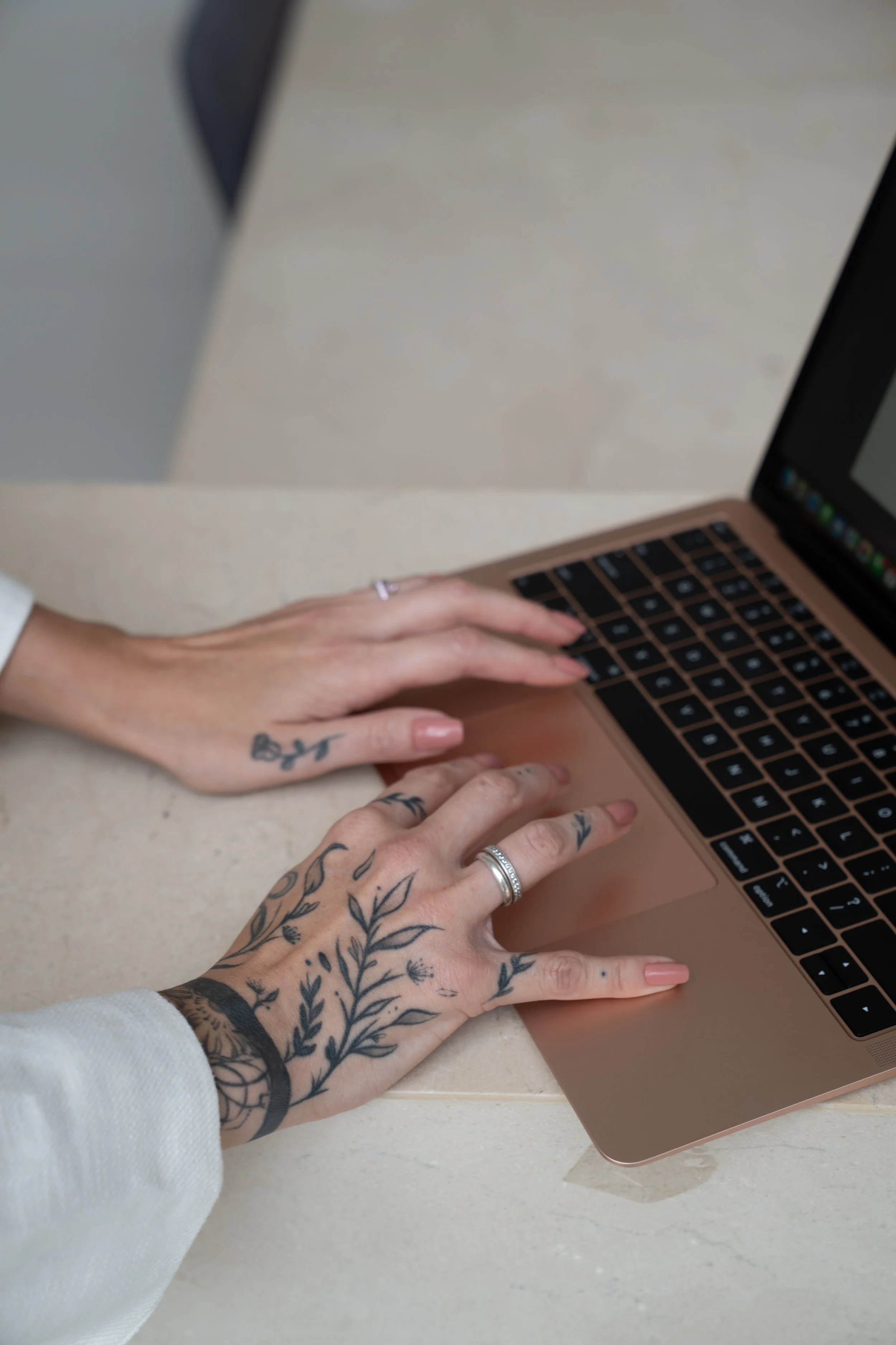 Mãos tatuadas usando um anel prateado, digitando em um laptop rosa dourado sobre uma mesa de mármore