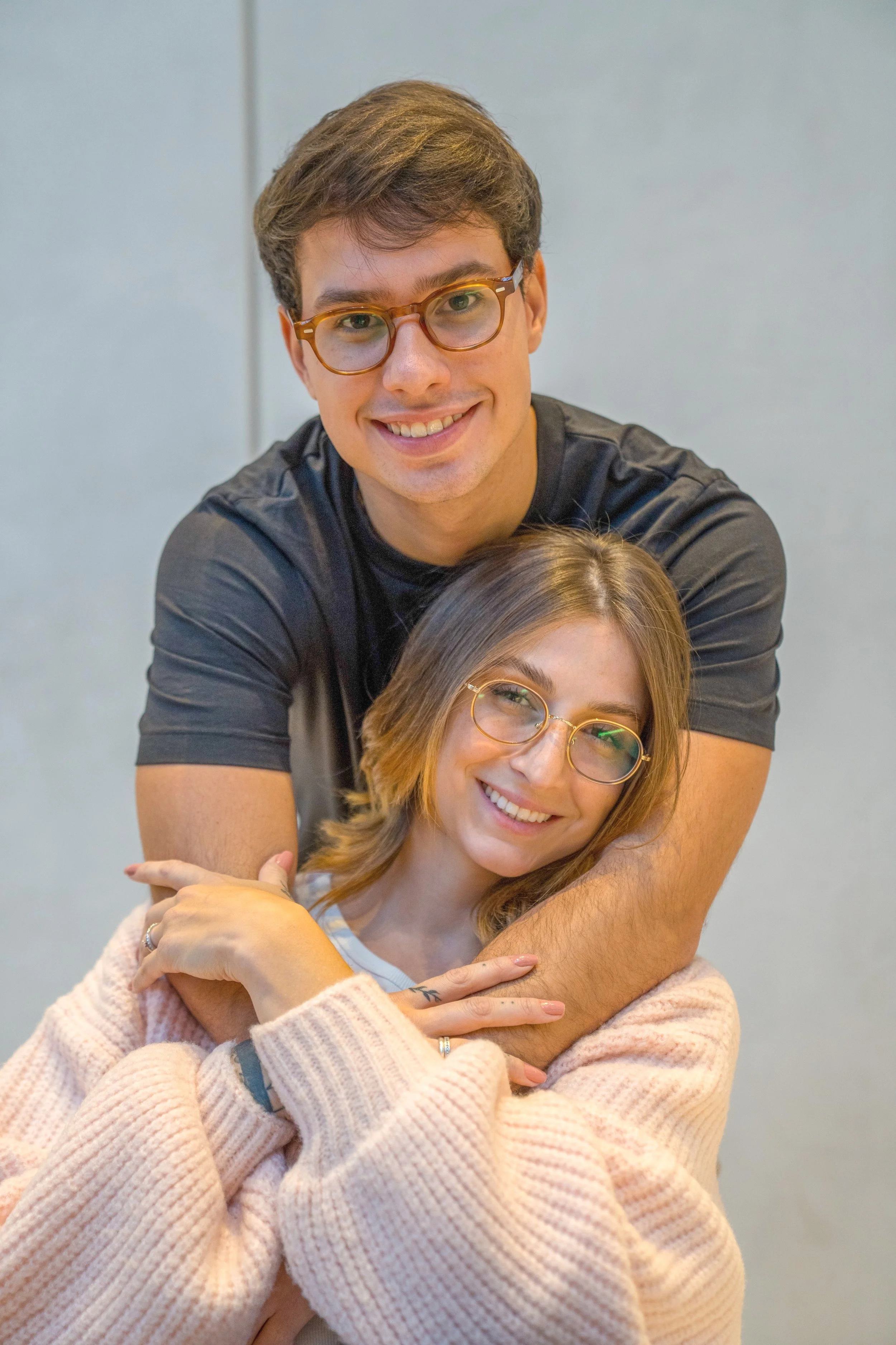Homem e mulher sorrindo, com o homem abraçando a mulher por trás, ambos usando óculos, em uma pose aconchegante e feliz.