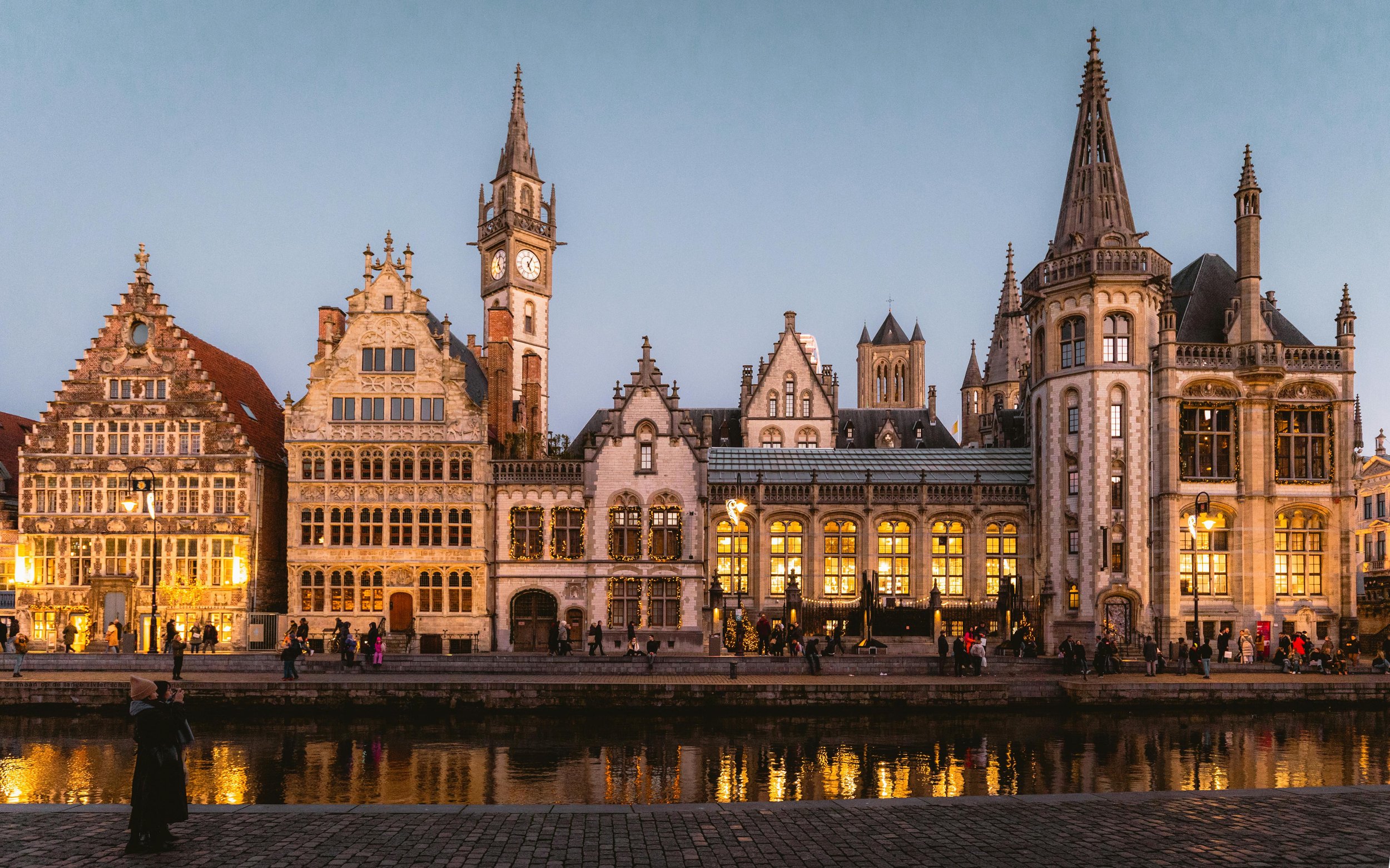 A canal-side shot of Ghent, with night time fast approaching. The shot is dominated by medieval buildings, some of which are lit up by interior lighting. People mill about in front of the buildings, with a canal running across the foreground
