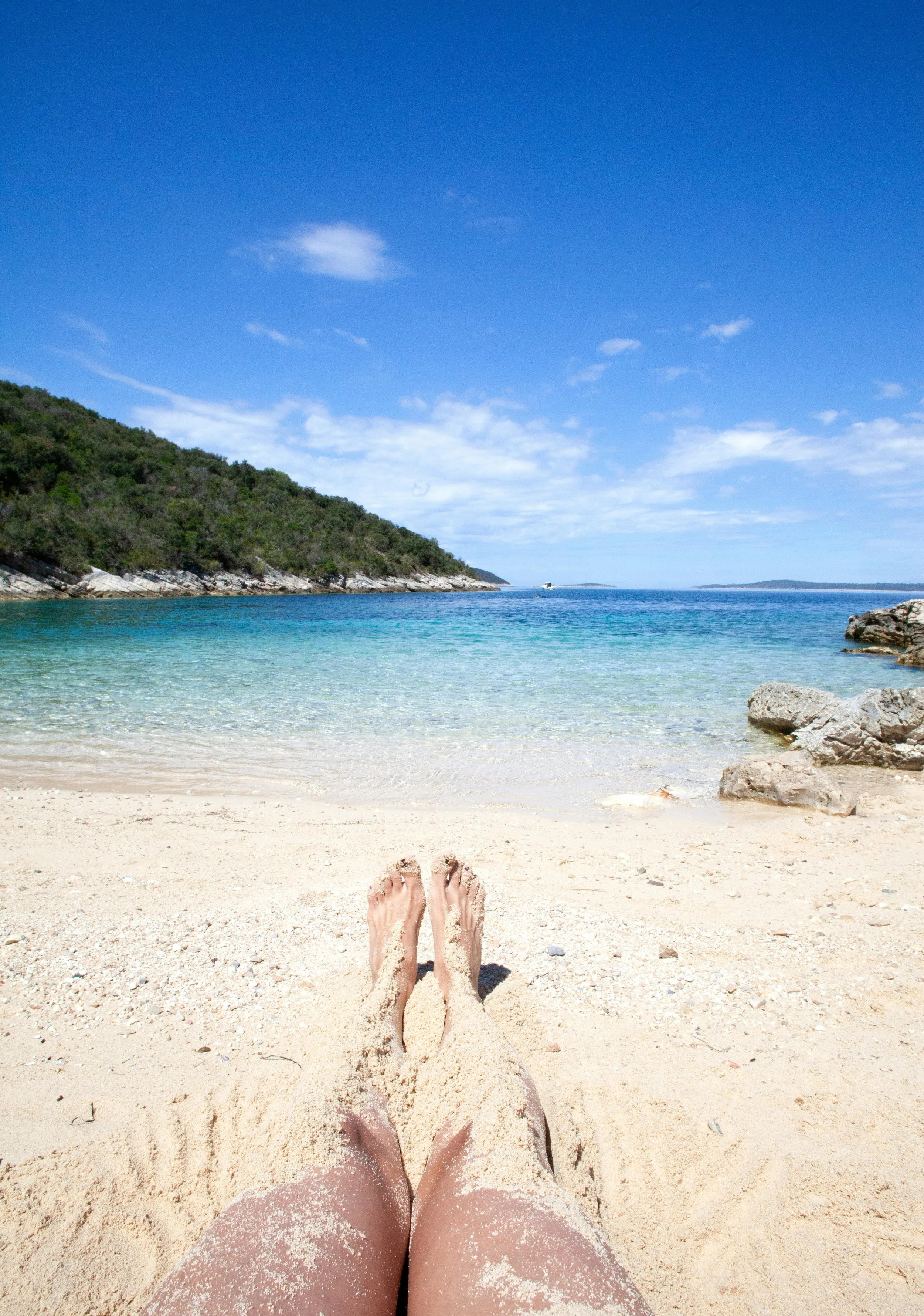 Two legs covered in white sand sit at the bottom of shot. Beyond is a clear blue ocean, green hills and a deep blue sky with whisps of white cloud.