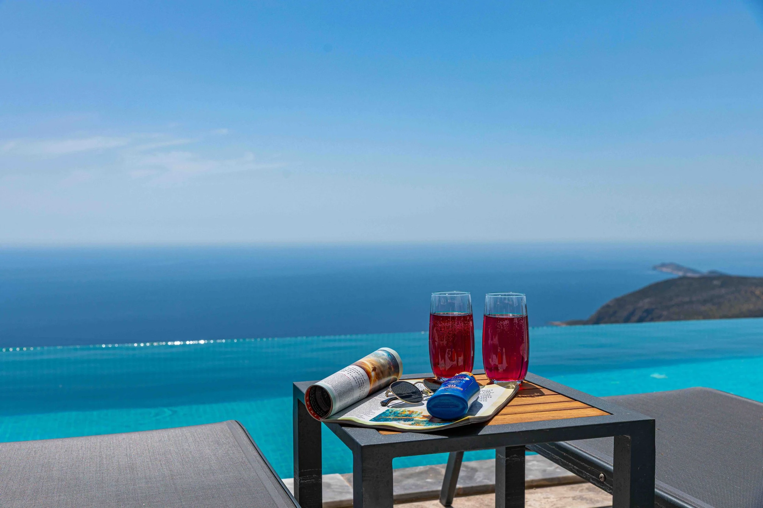 Two cocktails sit on a small side table, with a sun lounger on either side. Beyond is an infinity pool looking out to an ocean stretching to the horizon