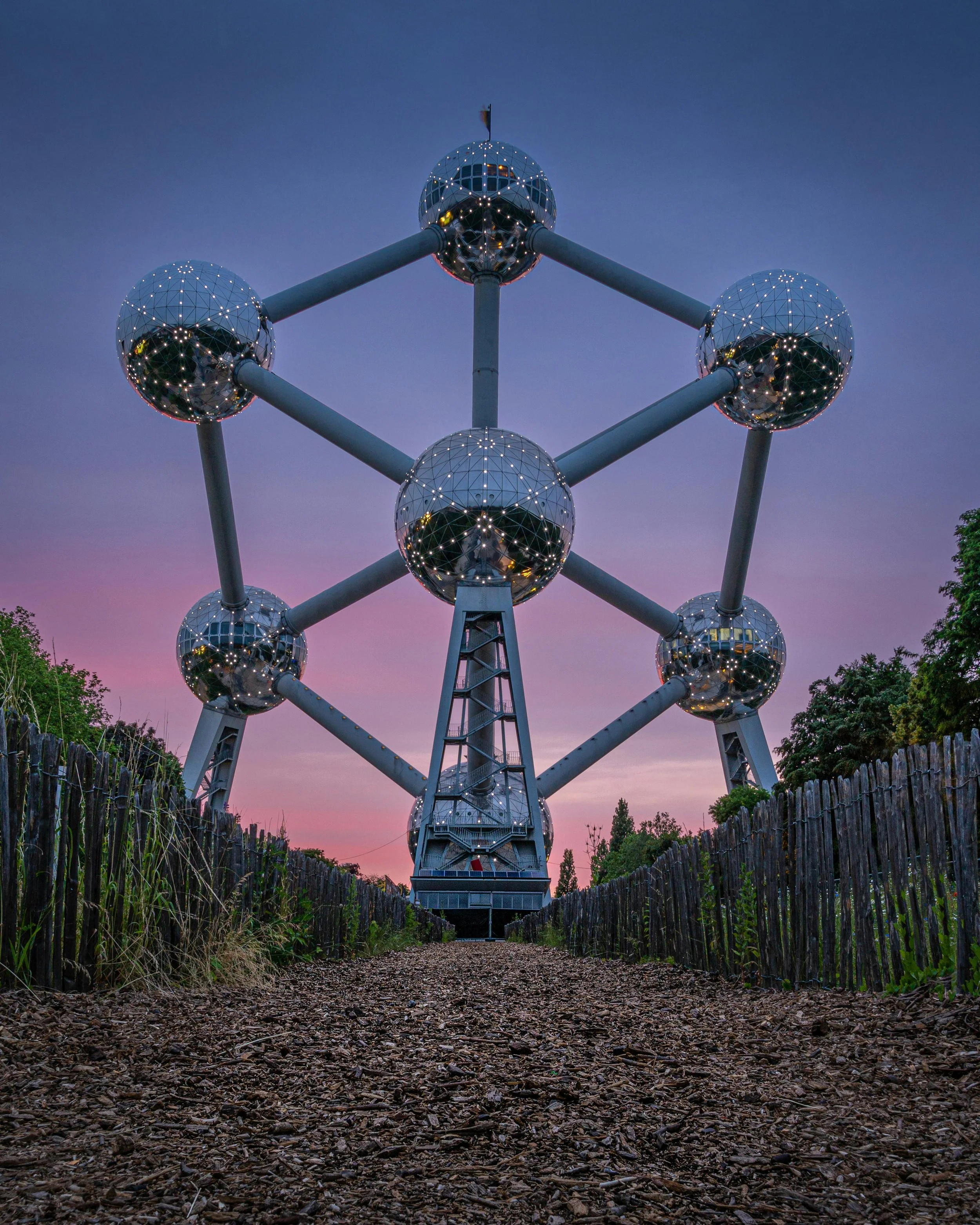 The camera is angled upwards, with the frame dominated by the Atomium, a famous landmark in Brussels featuring huge spheres connected by spindles. It's dusk, with a purple sky. Each sphere is lit with individual/multiople lights.