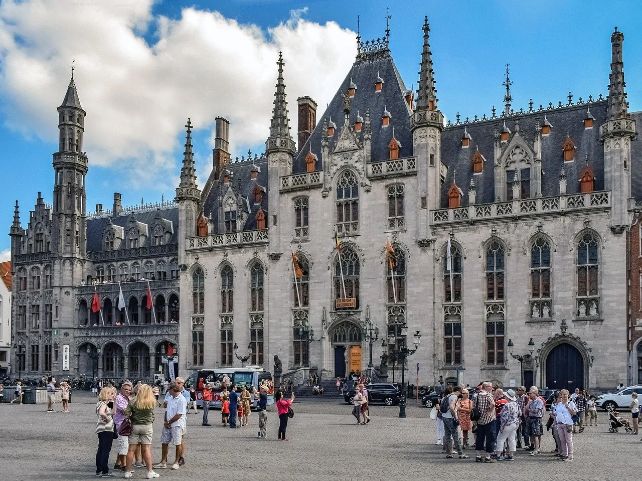 Daytime shot of the market square, with impressive period archtiecture in the background. People are gathered in groups in the square. A cloudy blue sky lines the top of the shot