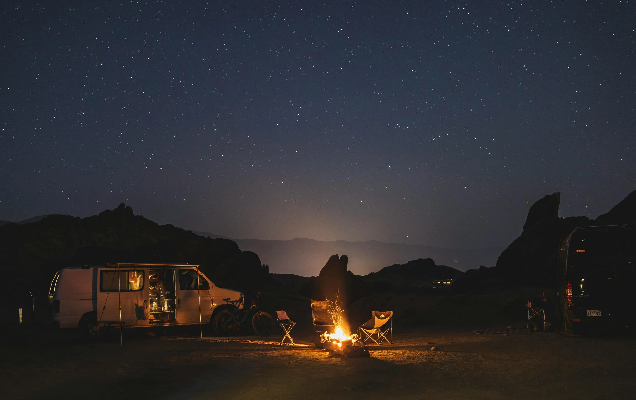 The shot is dominated by a dark starlit sky. Silhouettes of hills can be seen beyond a campfire which sits centre of frame. There are 3 chairs around the fire, and a campervan with awning attached sat beyond and to the left