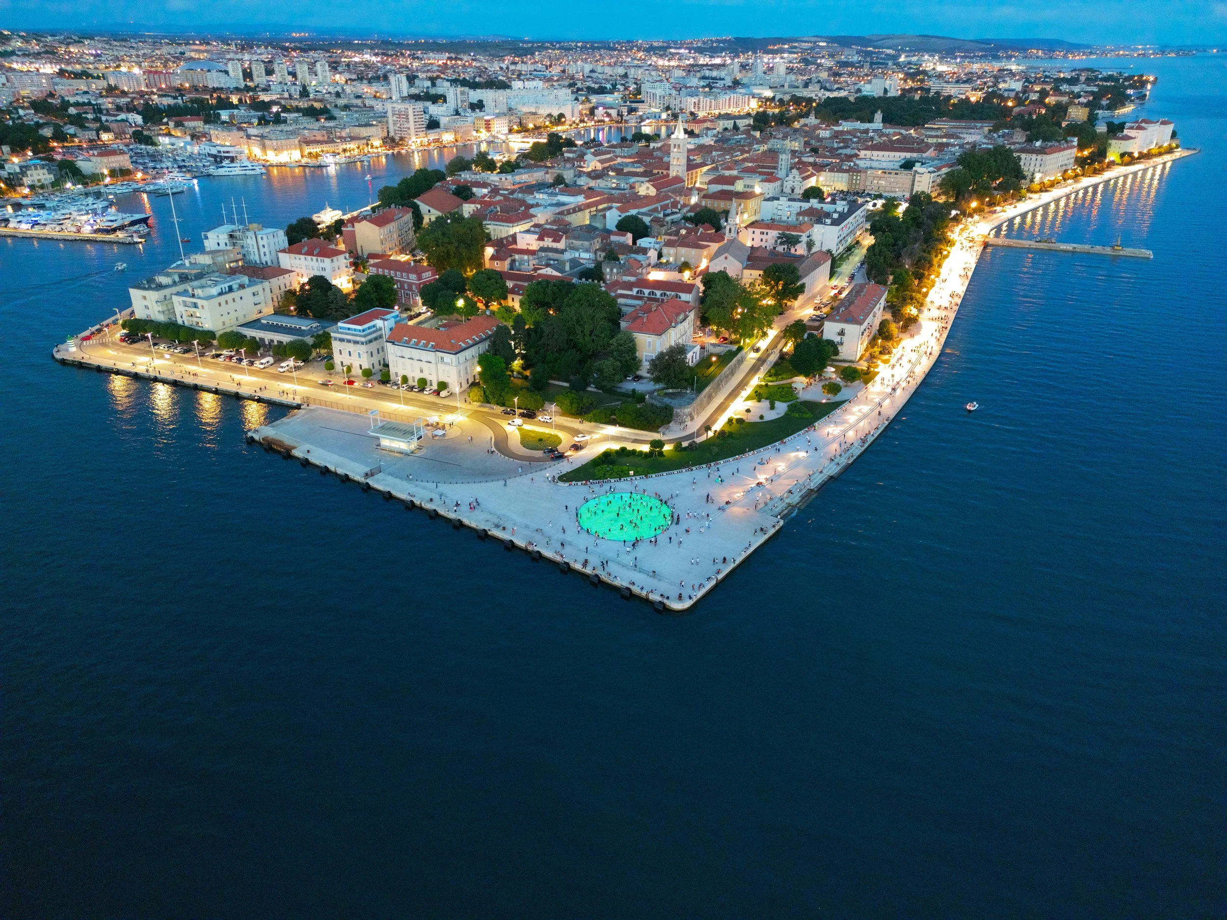 An earial view of Zadar by night. The angular peninsula is lit by street lights, surrounded by dark blue water. On the mainland, a mixture of white buildings and green trees sprawl across the city