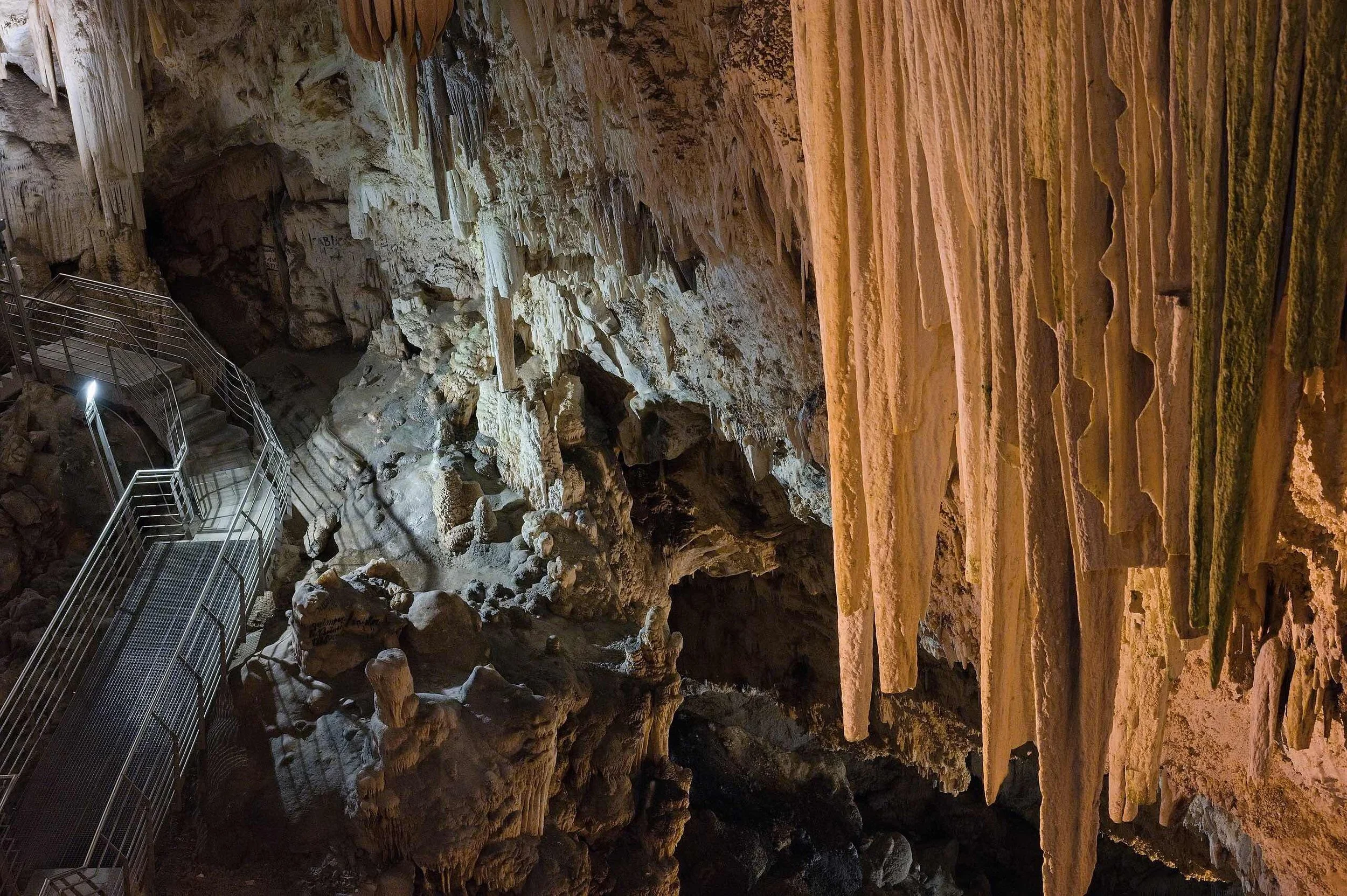Huge stalactites dominate right of frame, as we look down deep into a cave. A metal walkway can be seen below.