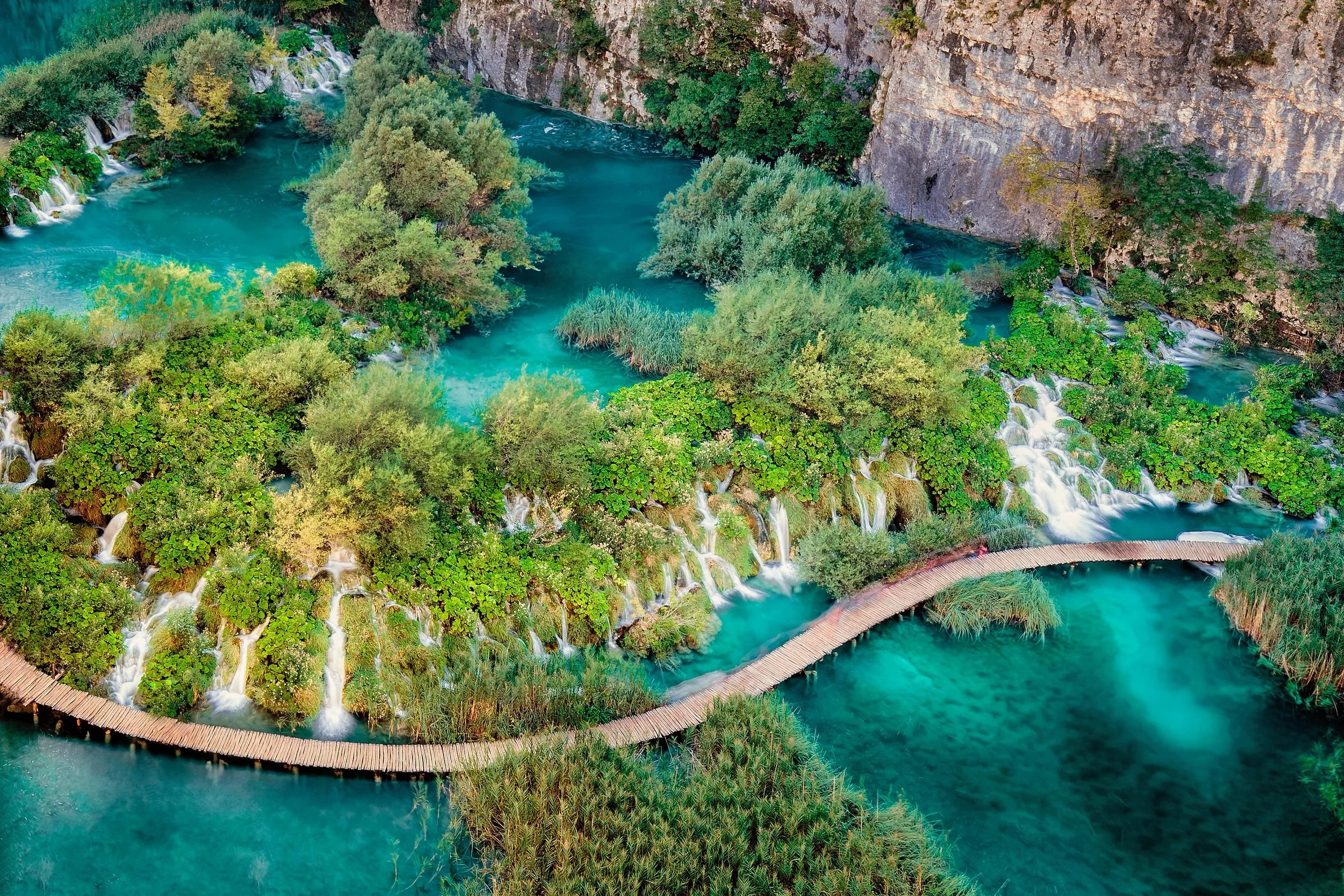 An aerial view of the turquoise waters of Plitvice lakes, with waterfalls splitting the greenery across the shot. A winding walkway bends across the water next to the waterfalls