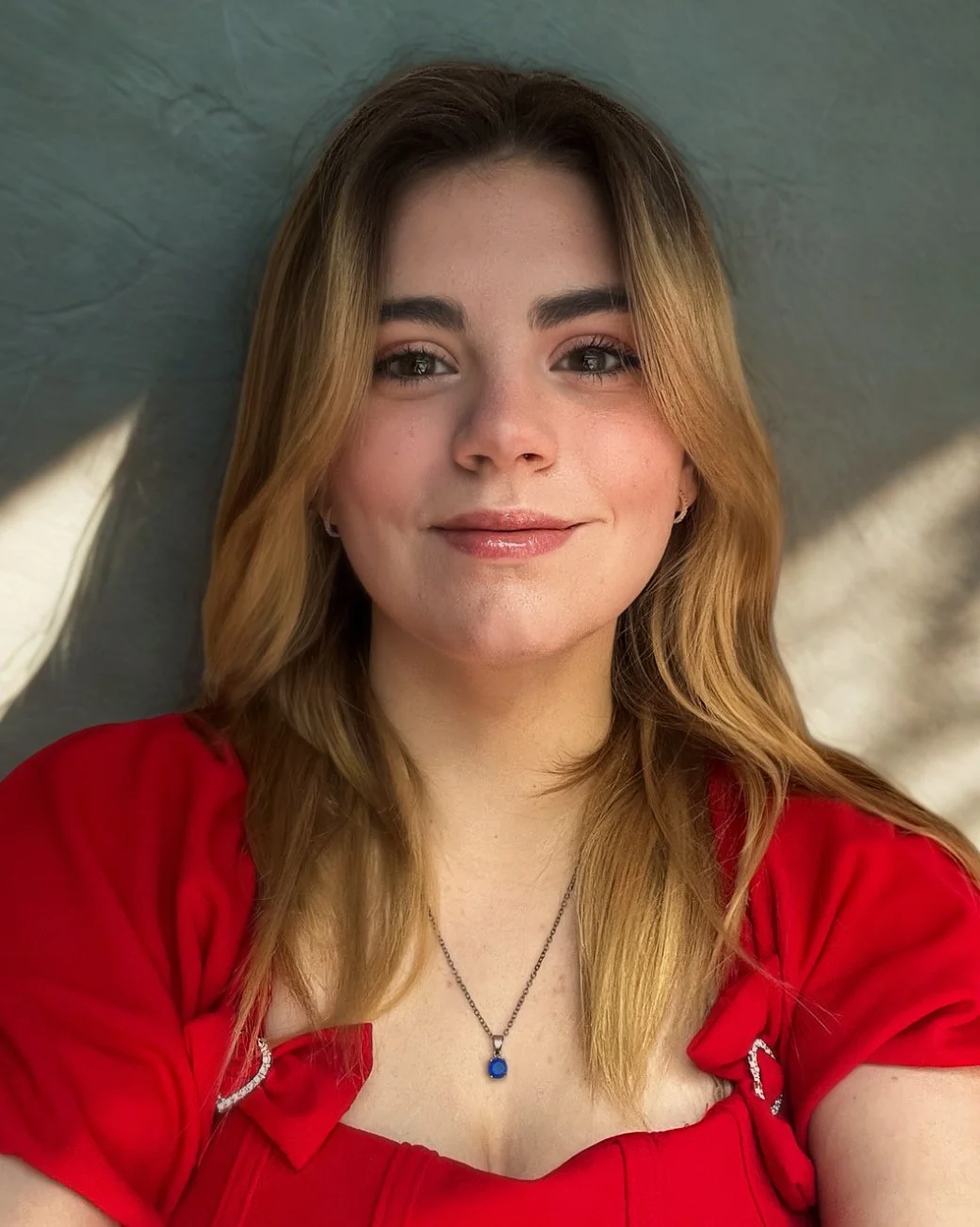 A young woman with long light brown hair smiling, wearing a red top and a silver necklace with a blue pendant, standing against a textured wall with soft lighting.