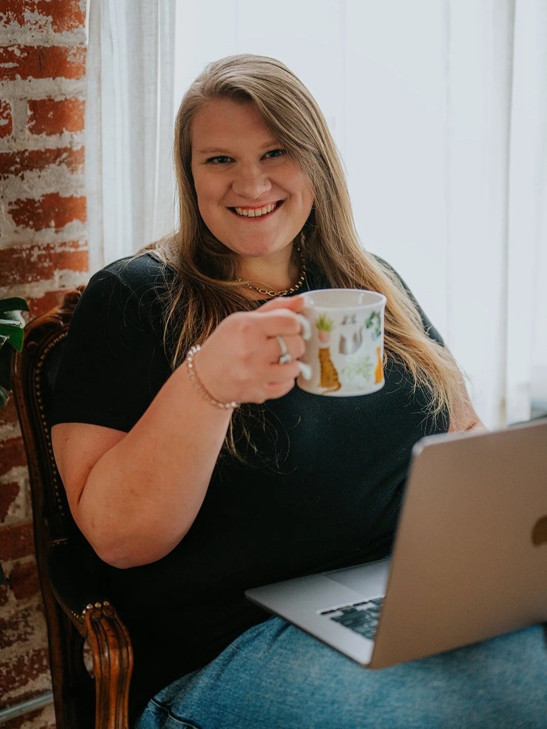 A smiling woman with long light brown hair holding a decorative mug, sitting in a room with a brick wall and a window, with a laptop in front of her.