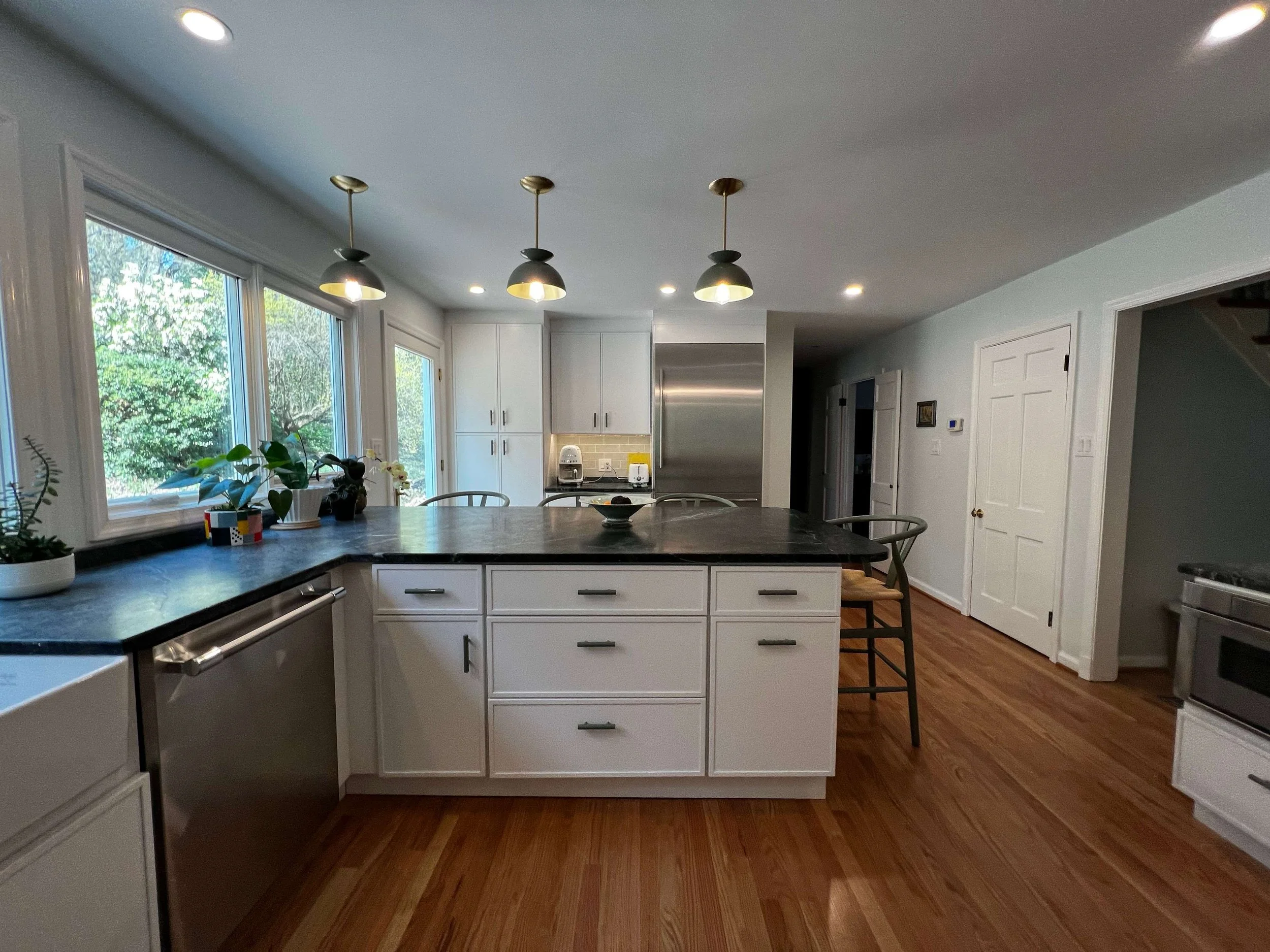 Modern kitchen with white cabinets, black countertop island, stainless steel appliances, hardwood floors, large windows with greenery outside, and pendant lights overhead.